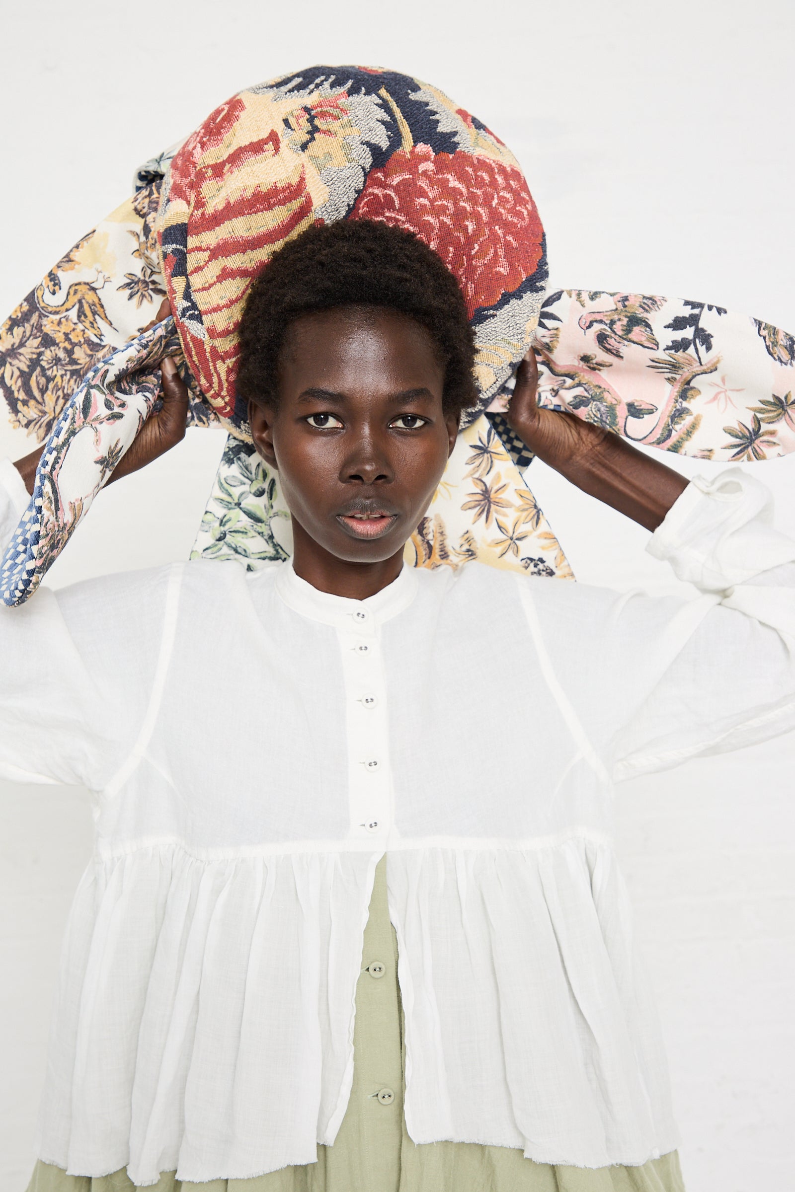 A person with short curly hair, wearing a white shirt, holds a colorful, patterned fabric above their head against a plain white background, resembling the intricate designs of a Wiener Times Cushion #2409 Rene Magrite Lisieux Tapestry.