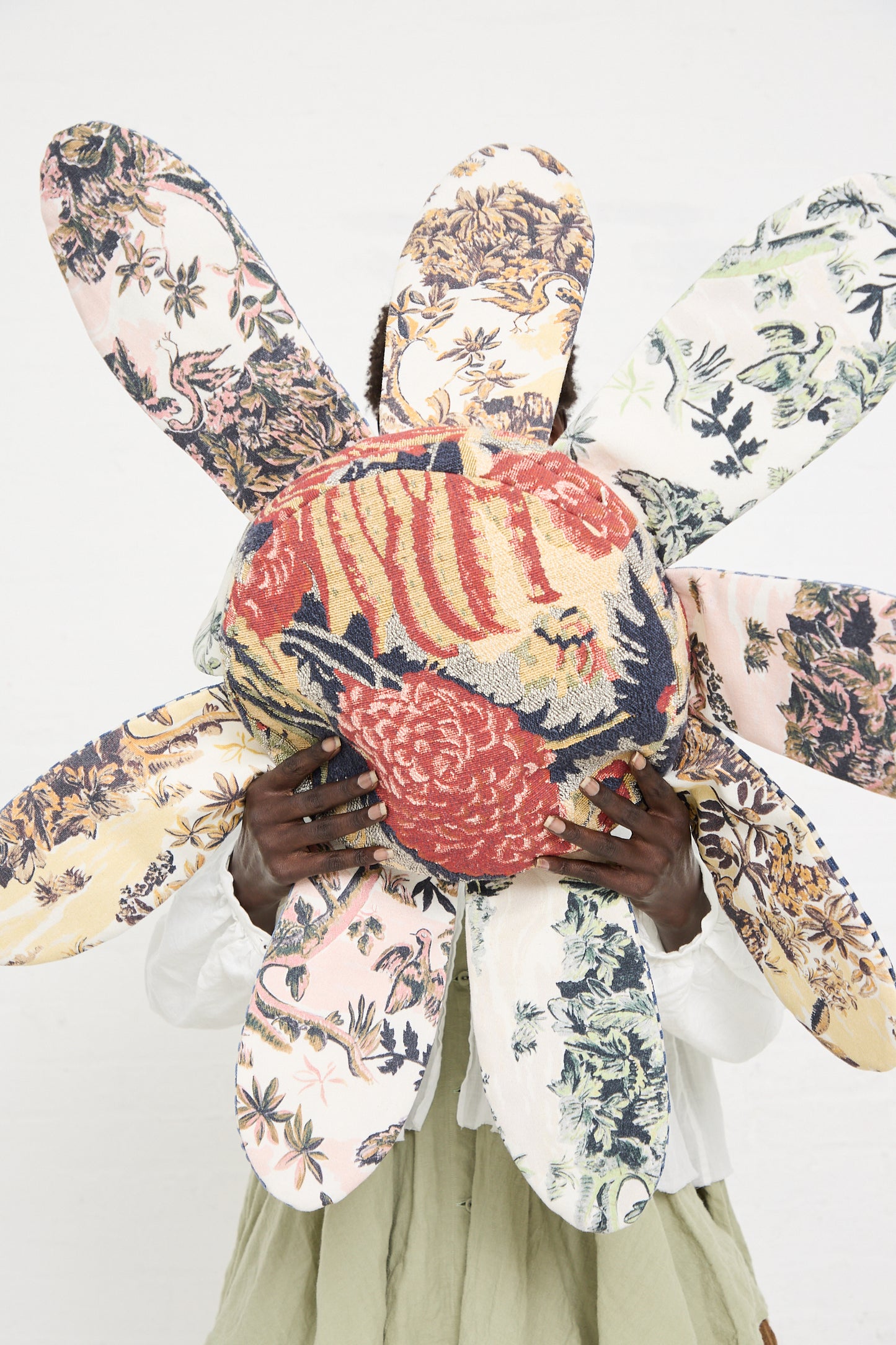 Person holding a large, colorful, floral-patterned fabric art piece shaped like a flower, obscuring their face. This exquisite item resembles a Wiener Times Cushion #2409 Rene Magrite Lisieux Tapestry and brings an artistic touch to the plain white background.