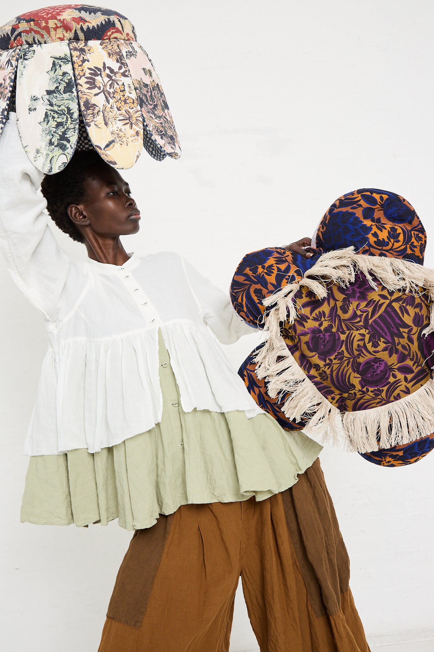 A person holding two large floral-patterned, flower-shaped cushions, wearing a layered white and green top with brown pants against a plain white background. One cushion resembles a Wiener Times Cushion #2409 Rene Magrite Lisieux Tapestry pillow made in Austria.