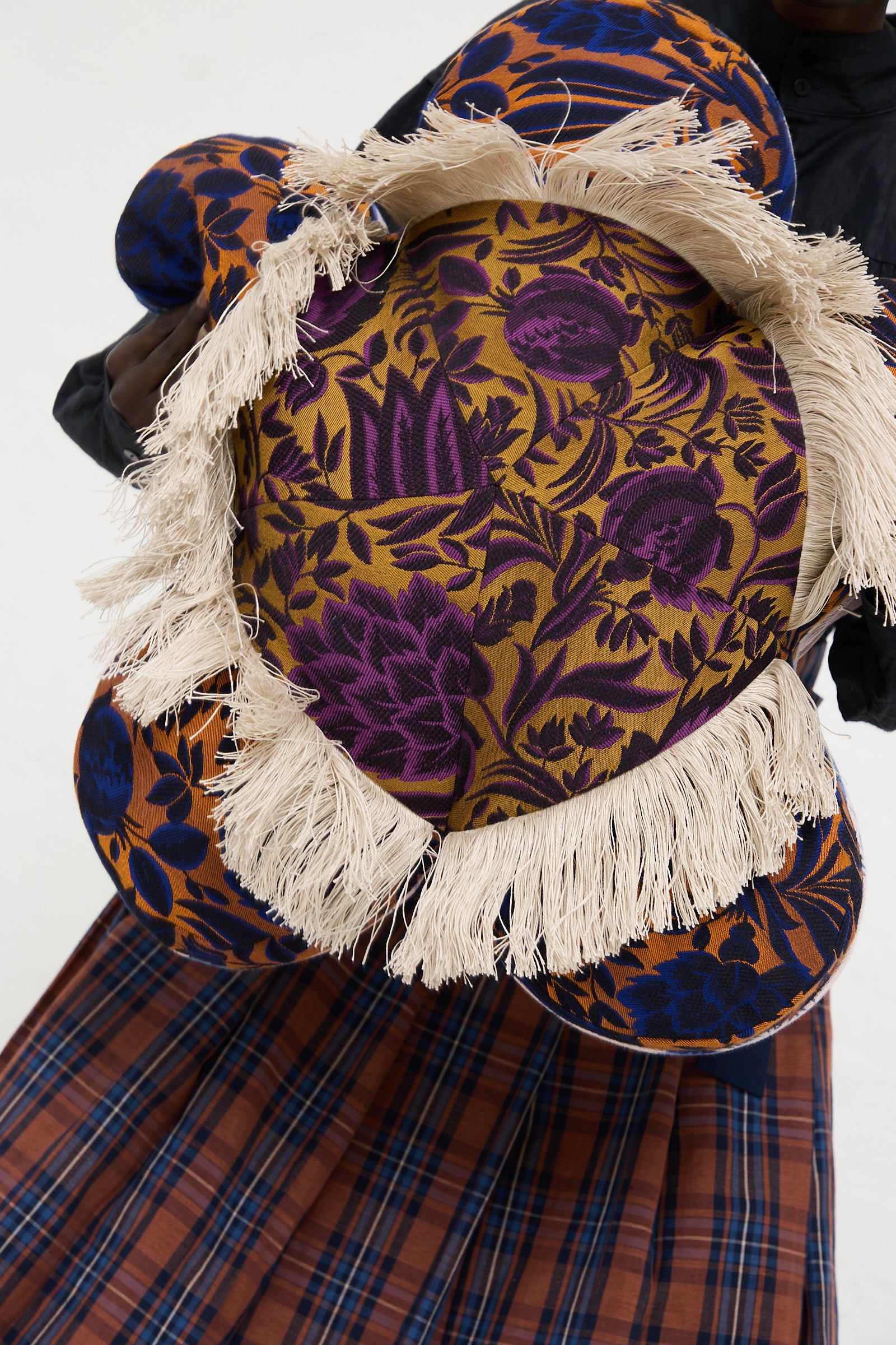 A person holds up a Cushion #2412 WW Jasmine Peche Passion by Wiener Times, adorned with a purple and gold floral pattern and beige fringe detail, wearing a dark top and a plaid skirt.