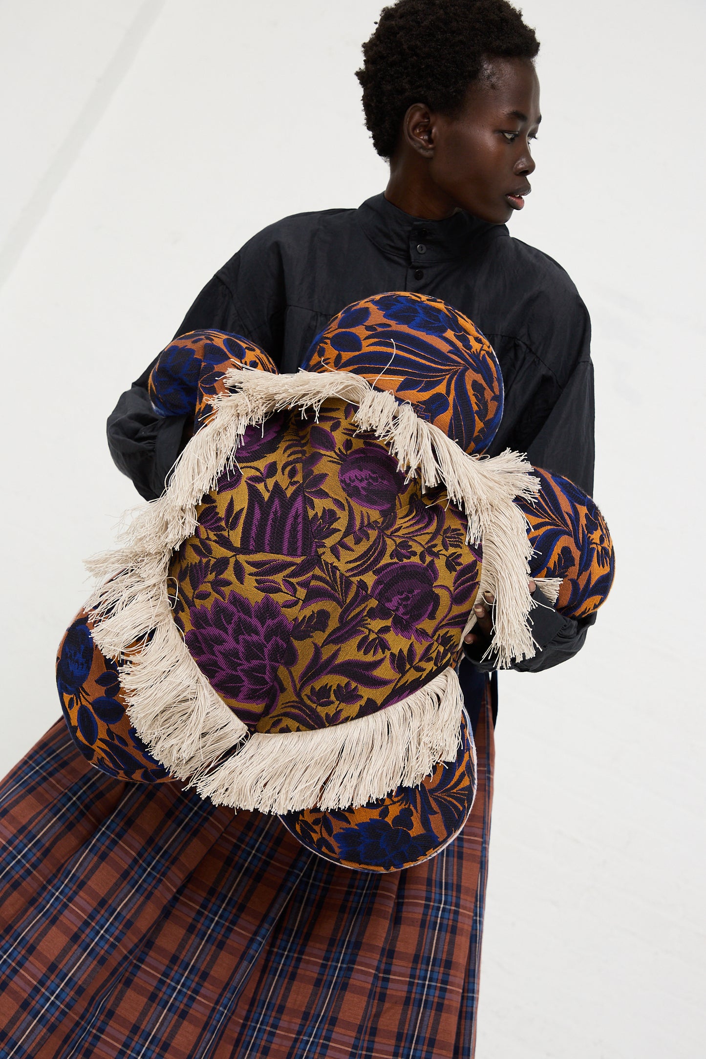 A person in a black shirt and plaid skirt holds a Cushion #2412 WW Jasmine Peche Passion by Wiener Times, adorned with a floral pattern and fringe detail, reminiscent of Wiener Werkstätte designs.