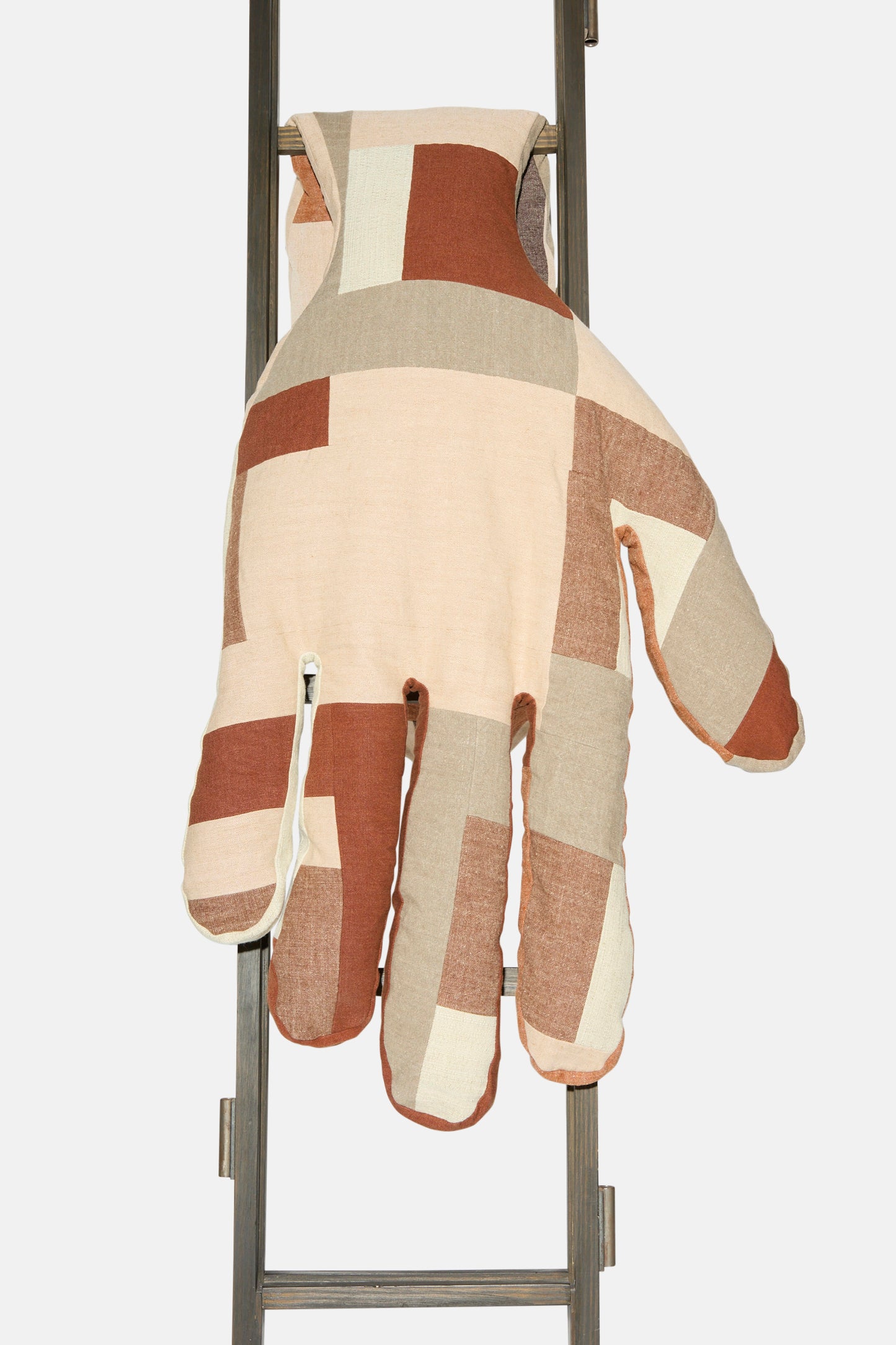 The Sonia Delaunay Tissu Simultané Giant Hand Bolster II by Wiener Times—a large, patchwork hand-shaped cushion in beige, brown, and cream—rests on a metal ladder, capturing the playful spirit of the Wiener Werkstätte.