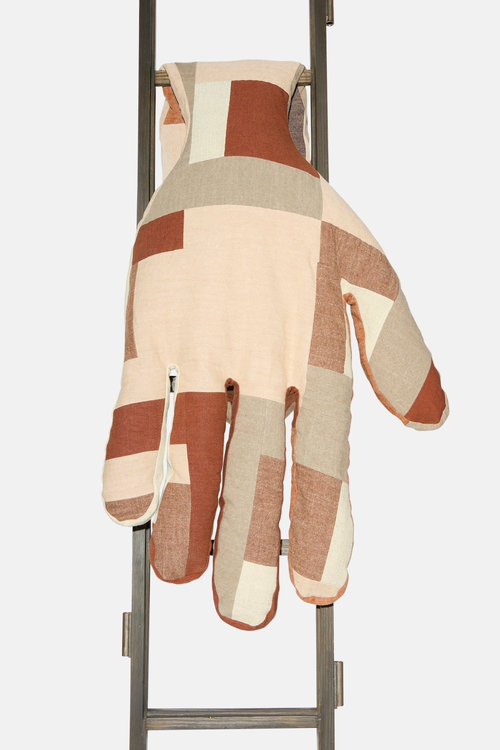 The Sonia Delaunay Tissu Simultané Giant Hand Bolster II by Wiener Times—a large, patchwork hand-shaped cushion in beige, brown, and cream—rests on a metal ladder, capturing the playful spirit of the Wiener Werkstätte.