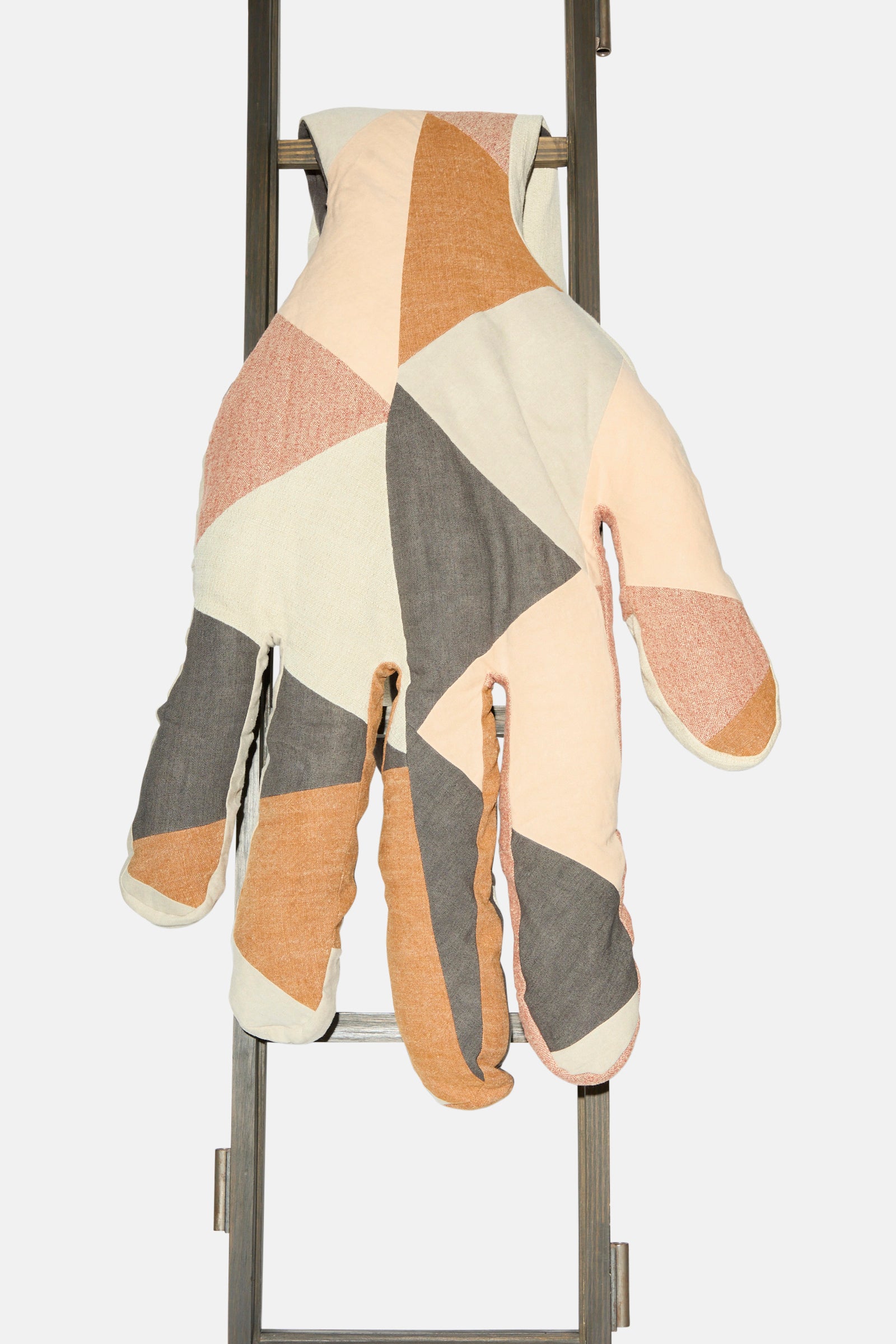 The Sonia Delaunay Tissu Simultané Giant Hand Bolster I by Wiener Times—a large, patchwork hand-shaped pillow inspired by Wiener Werkstätte—drapes over a metal ladder against a plain white background.