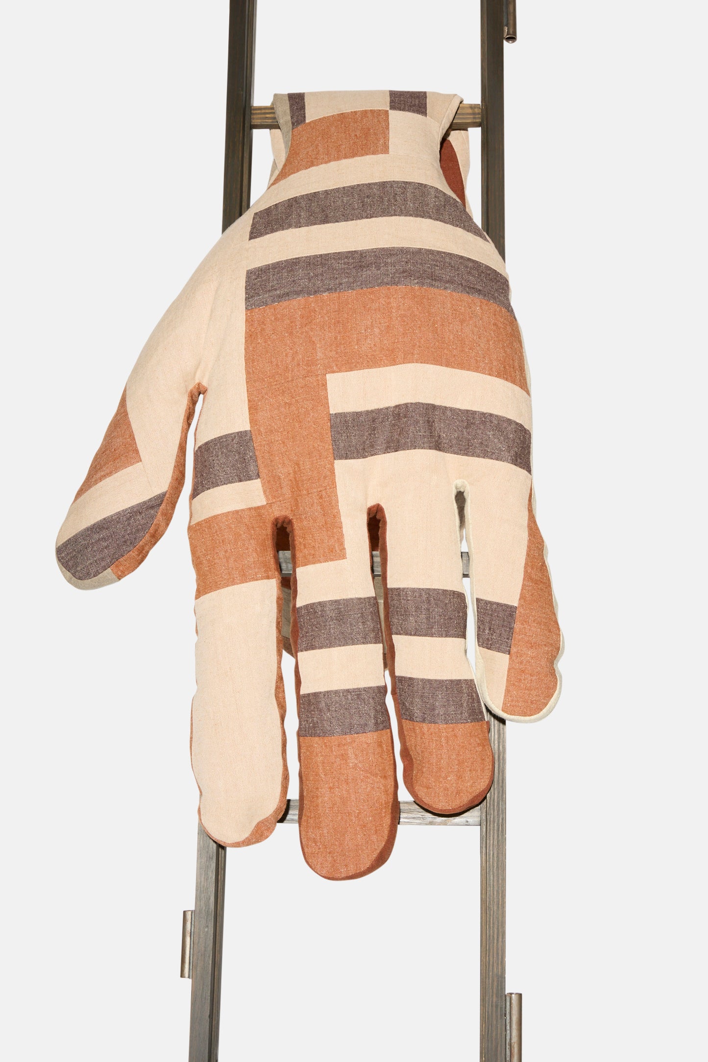 The Sonia Delaunay Tissu Simultané Giant Hand Bolster II by Wiener Times, featuring beige, brown, and tan stripes, rests over a metal ladder rung against a white background, reflecting Wiener Werkstätte-inspired artisanal design.