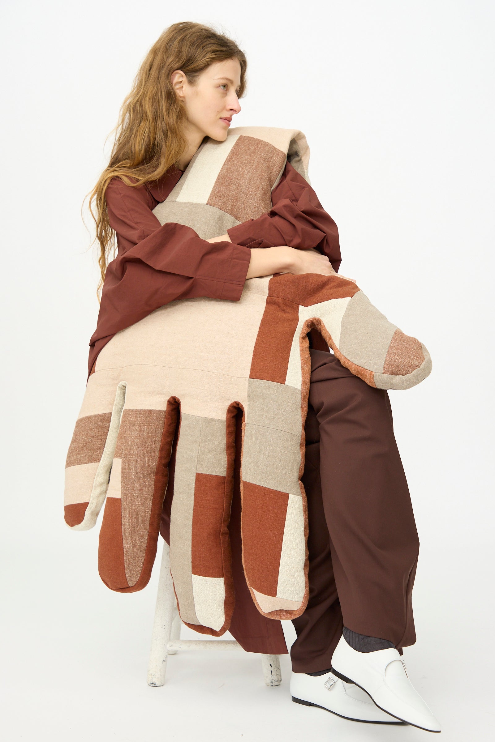 A woman in brown sits on a stool, holding the Wiener Times Sonia Delaunay Tissu Simultané Giant Hand Bolster II against a plain white background.