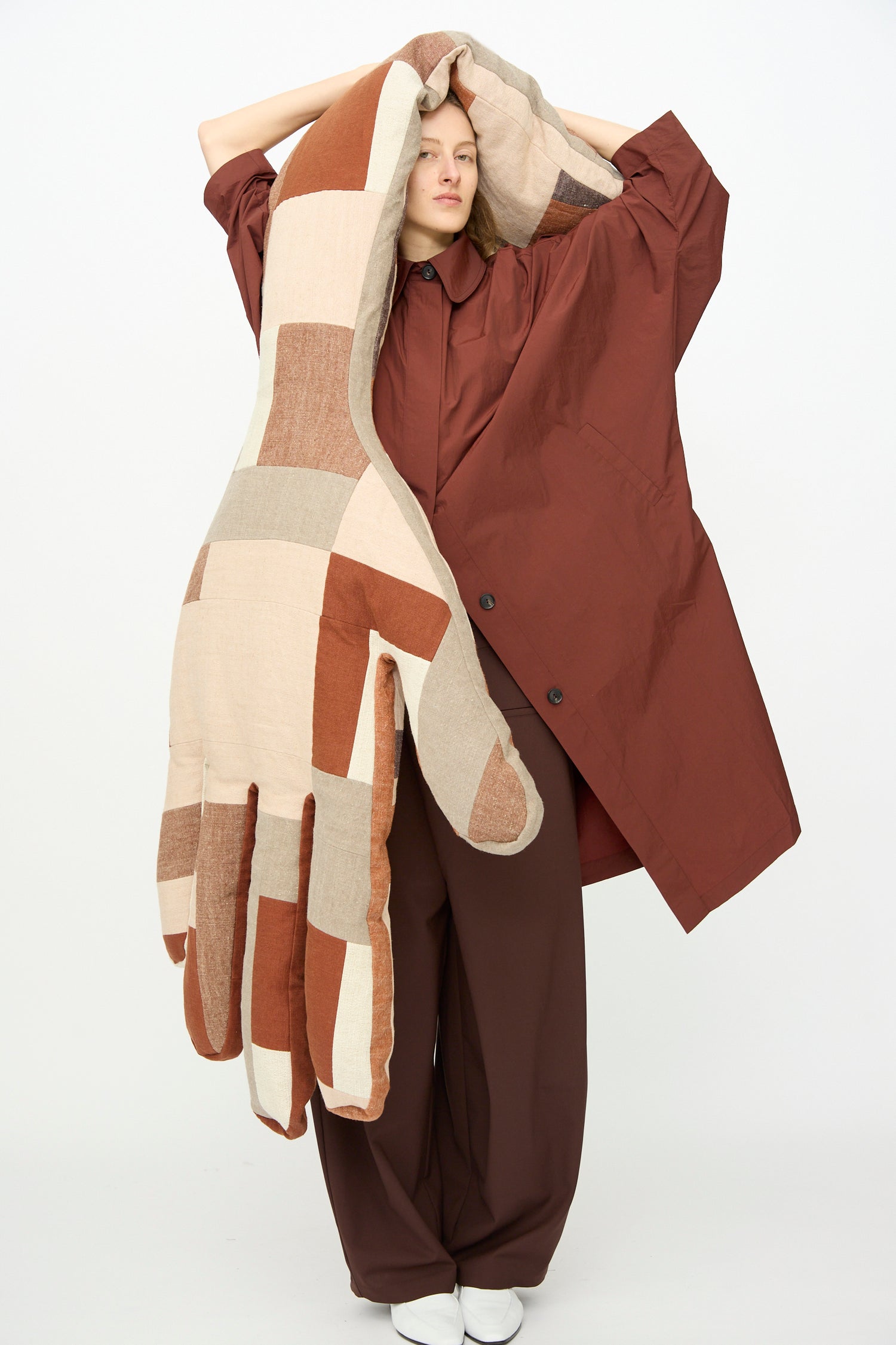 A woman holding up the Wiener Times Sonia Delaunay Giant Hand Bolster, wearing Corder's Batwing Trench in Sienna and Tailored Crossover Pant in Burgundy, against a white backgroud.
