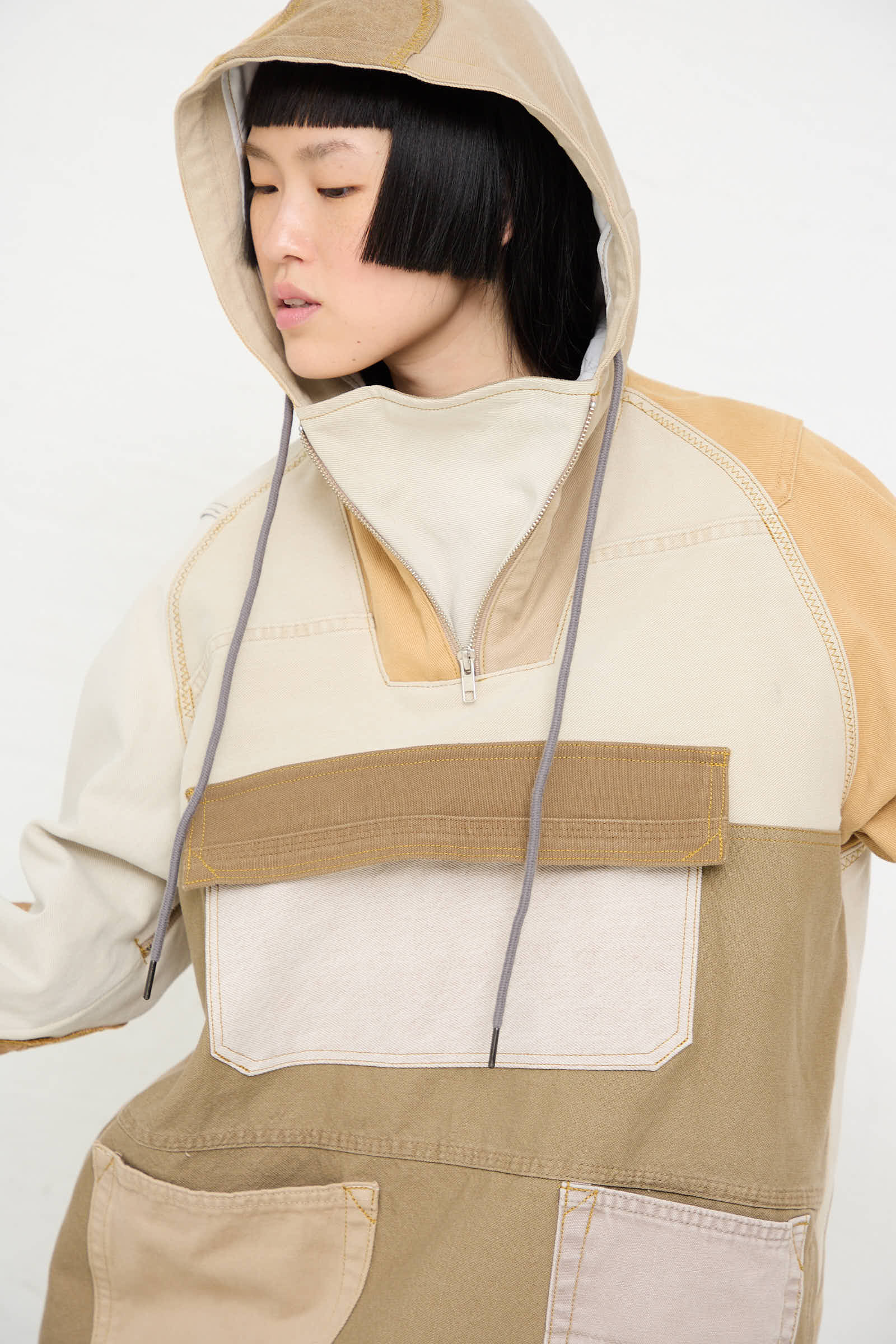 A woman with short black hair wears a handmade, one-of-a-kind Anorak in Beige and Brown by WildRootz, featuring a large front pocket. Crafted in New York, she gazes downward, with the hood loosely covering her head.