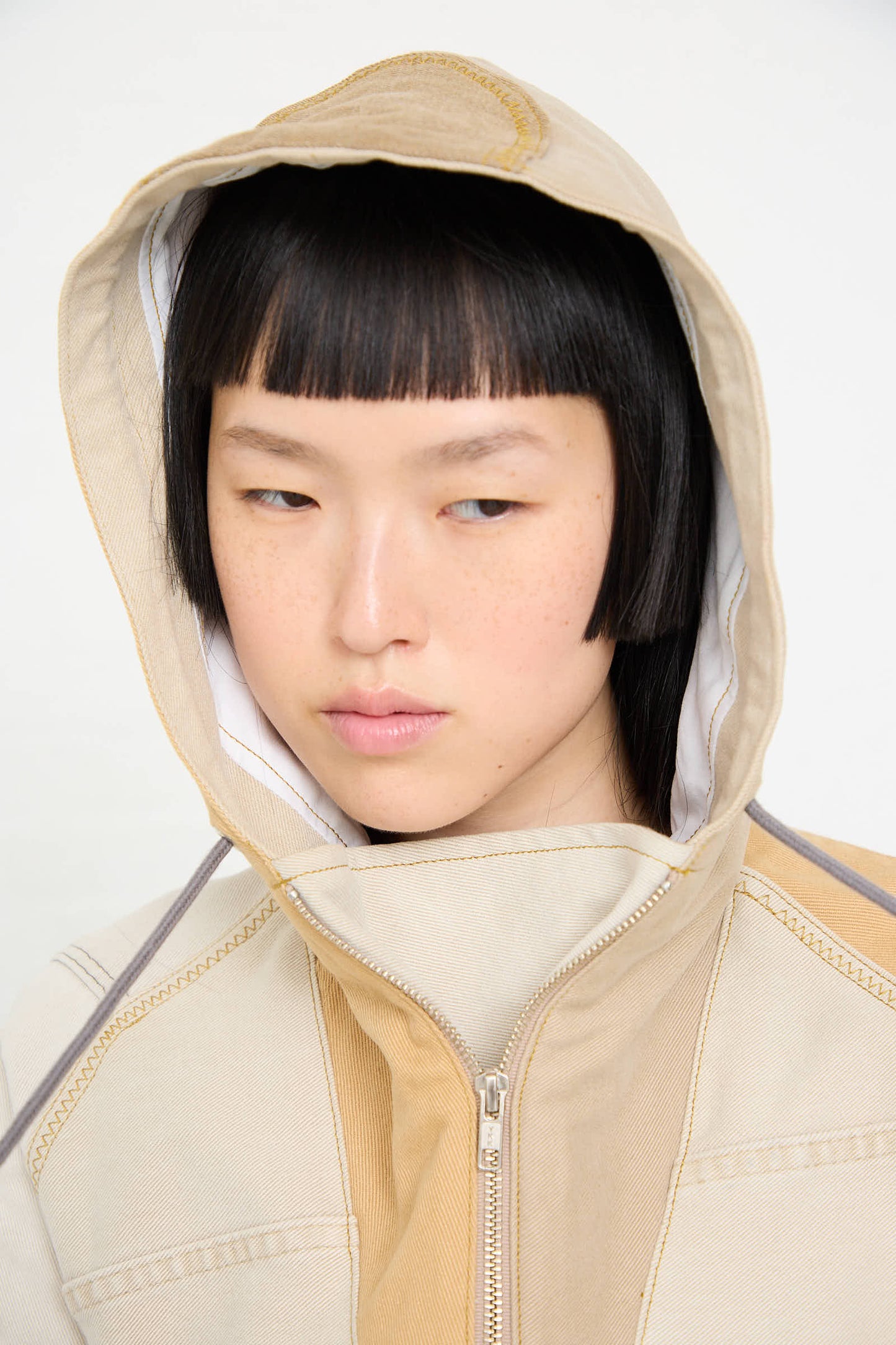 A person with short black hair wearing a handmade, one-of-a-kind Anorak in Beige and Brown by WildRootz looks downward with a neutral expression.