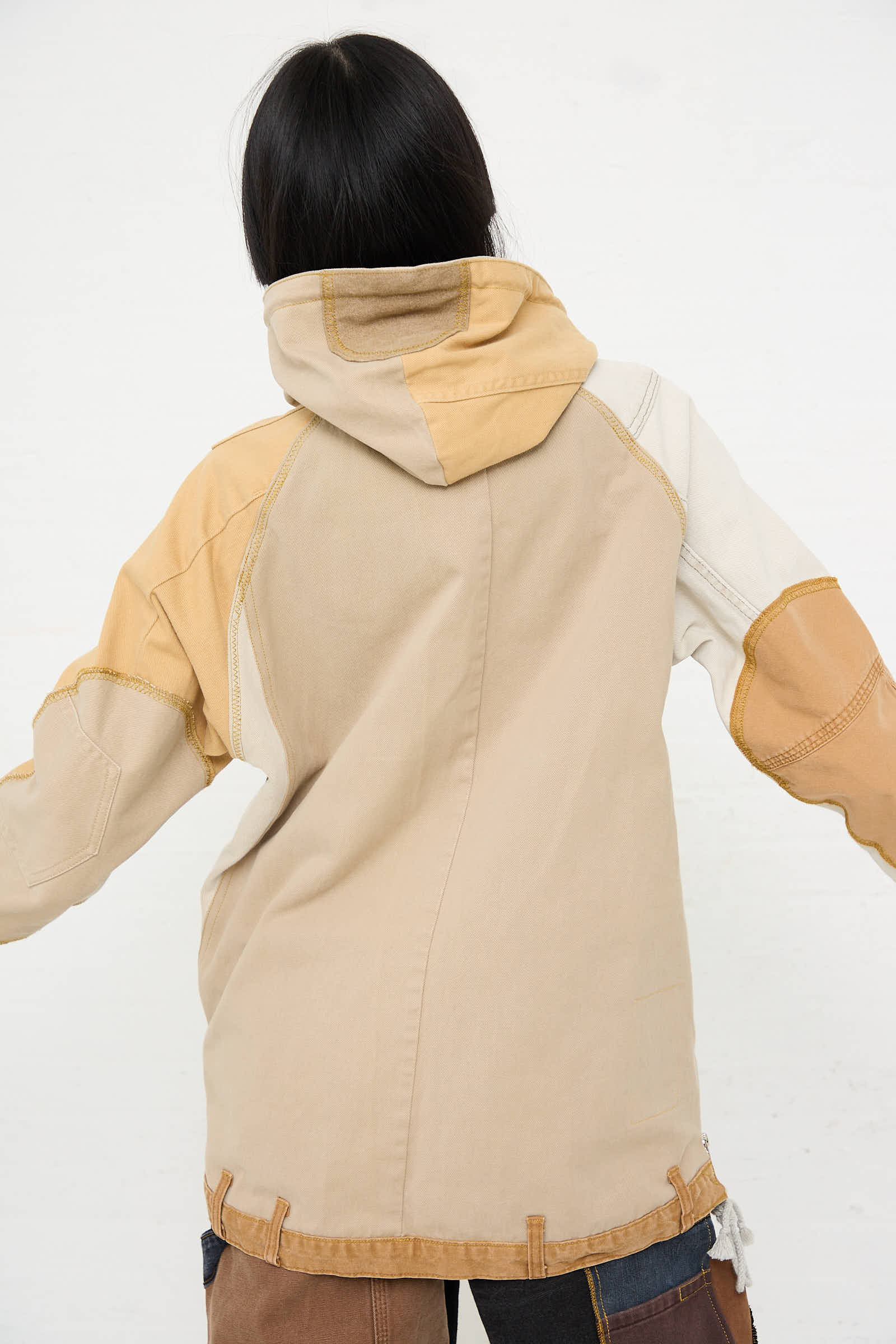 A person with long black hair faces away, donning the Anorak in Beige and Brown by WildRootz. This unique jacket made from recycled denim anorak materials showcases its color-blocked design in beige, tan, and white tones as they slightly outstretch their arms.