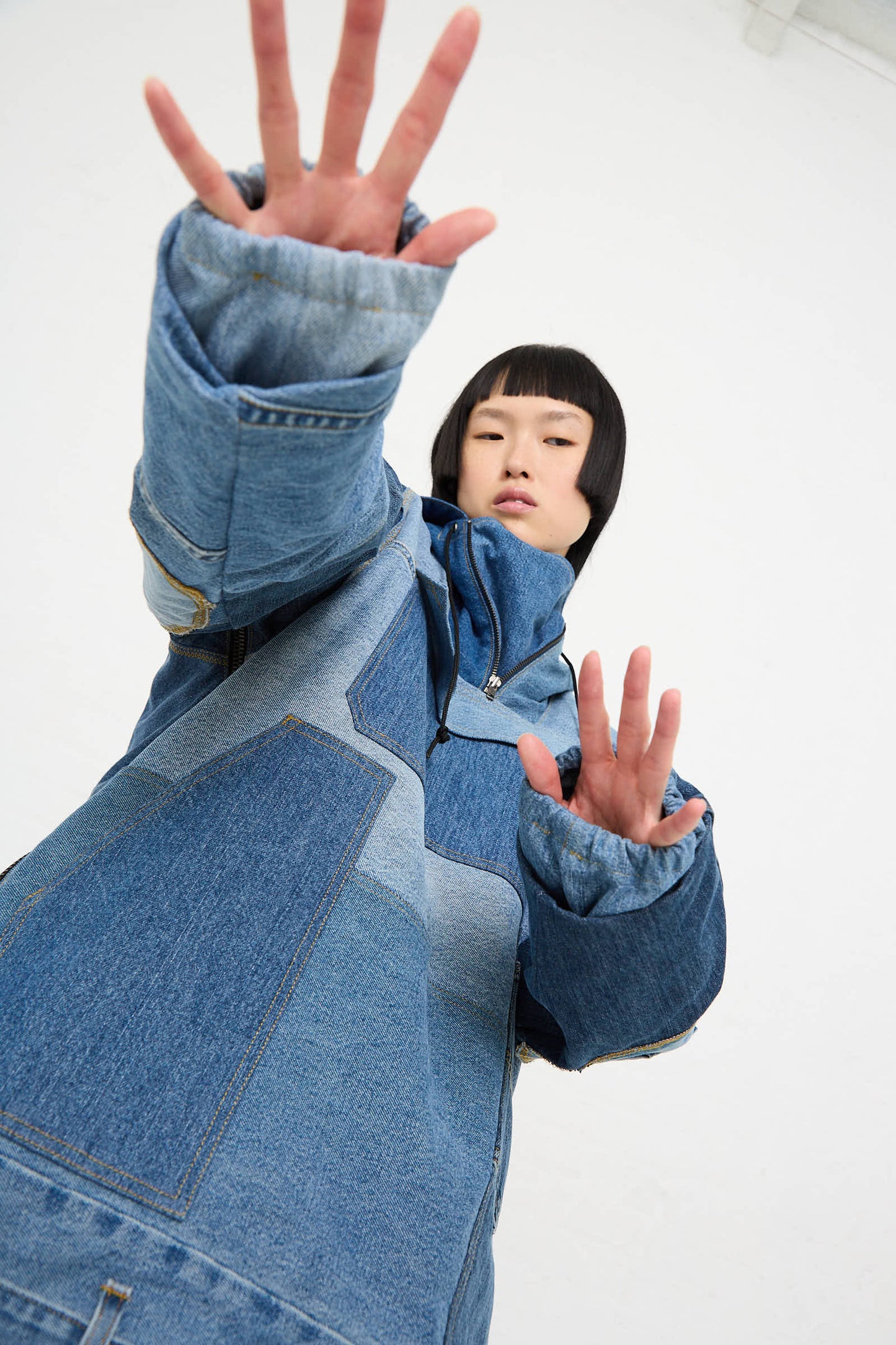 A person with short black hair wears a long Anorak in Blue by WildRootz made from recycled denim and extends their hands toward the camera against a plain white background.