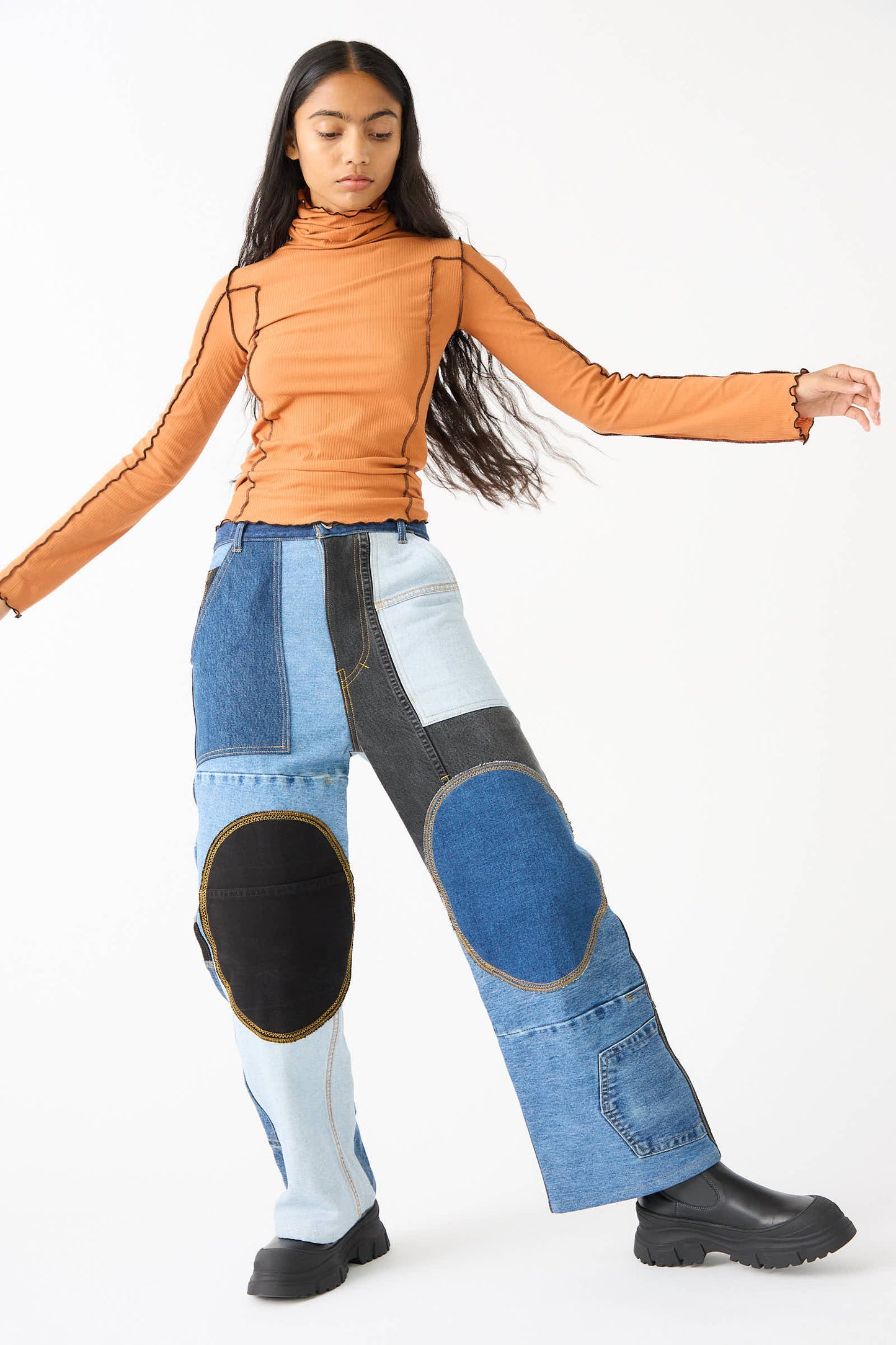 Person wearing WildRootz Reworked Jeans in Blue, Black, and Grey handmade in New York, an orange long-sleeve top, and black boots, standing against a plain white background.