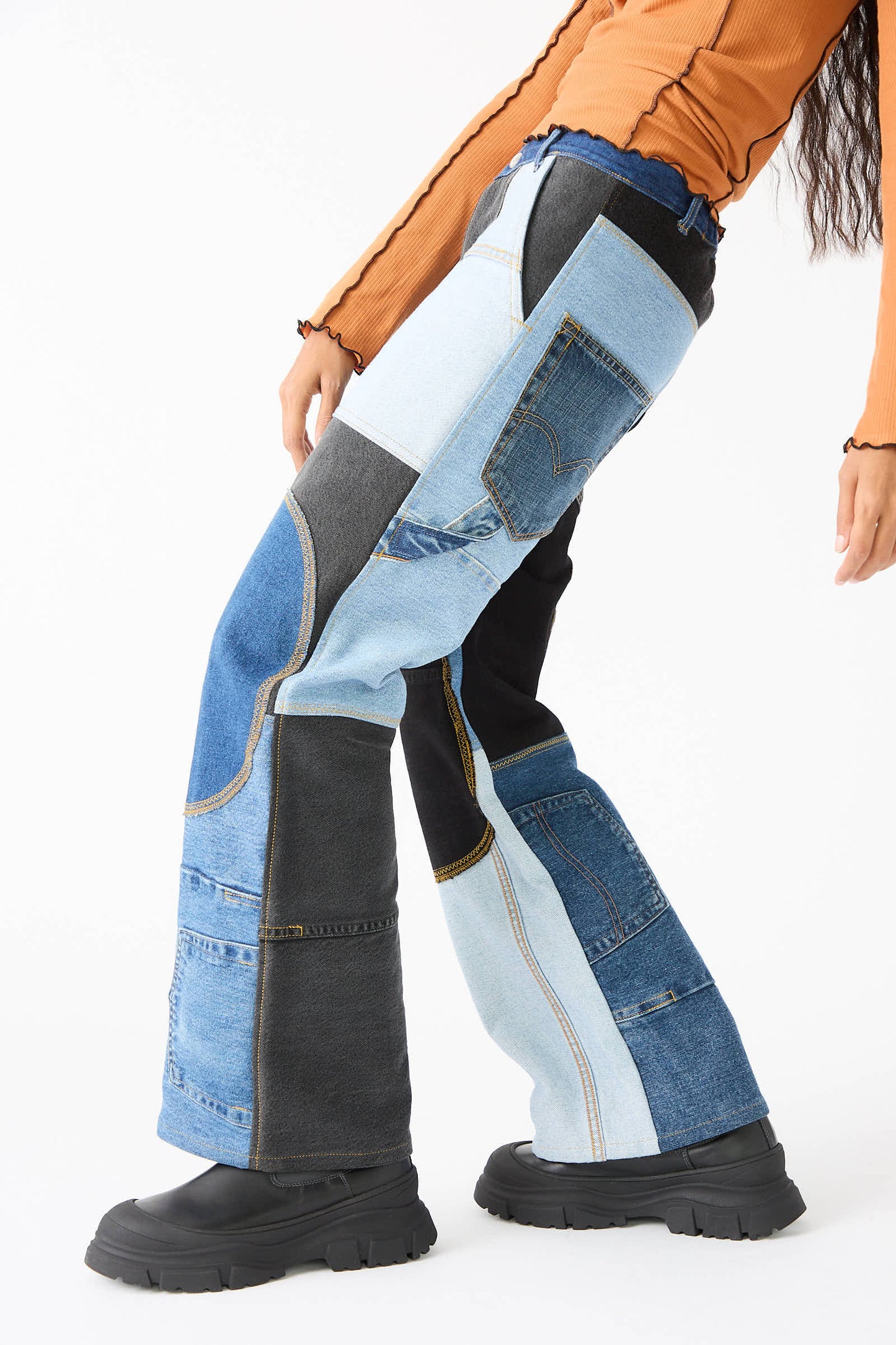 A person wearing WildRootz's Reworked Jeans in Blue, Black and Grey, handmade in New York, paired with an orange long-sleeve top and black shoes, stands against a white background.