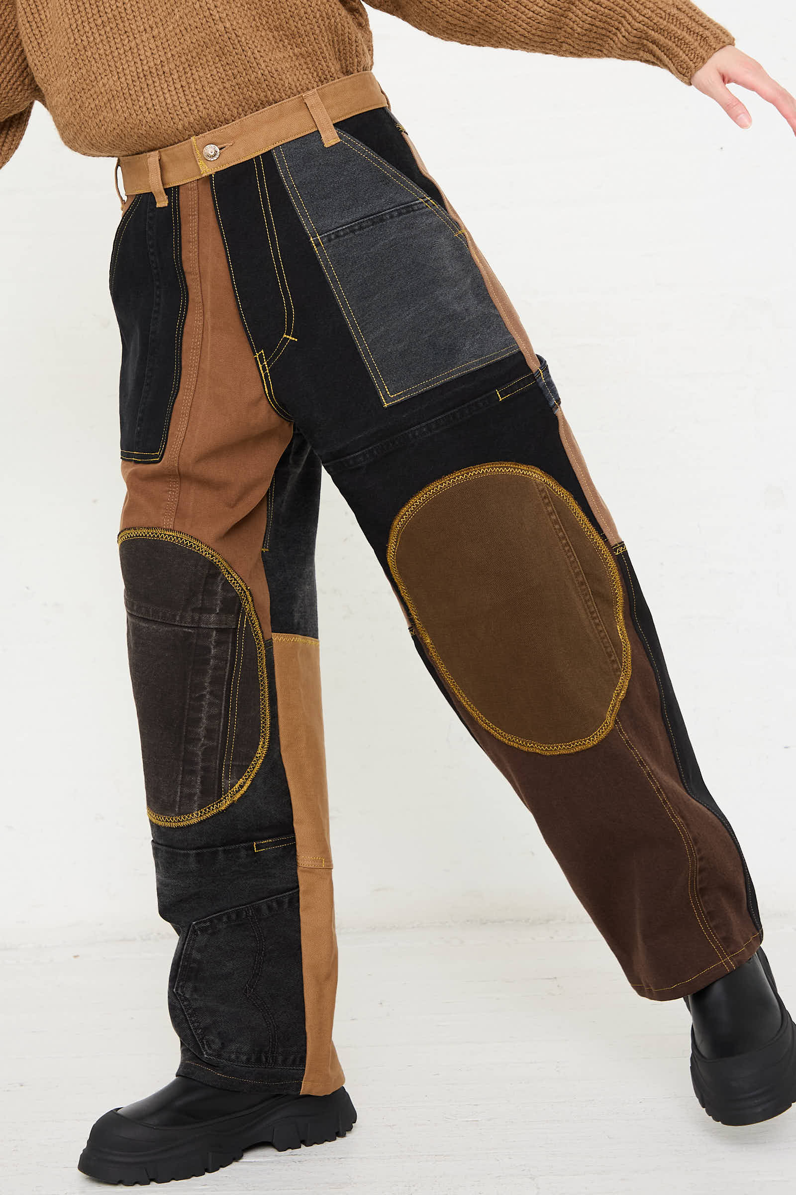 Person wearing WildRootz Reworked Jeans in Black and Brown I, handmade in New York from recycled denim with multiple colors and fabrics. These high-waisted beauties feature large knee patches and are paired perfectly with black boots and a brown sweater.