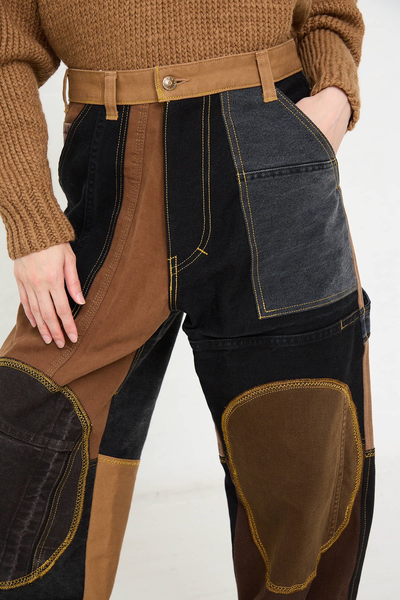 A person wearing WildRootz Reworked Jeans in Black and Brown I, handcrafted in New York, paired with a brown knit sweater. The person has one hand in the pocket of the jeans.