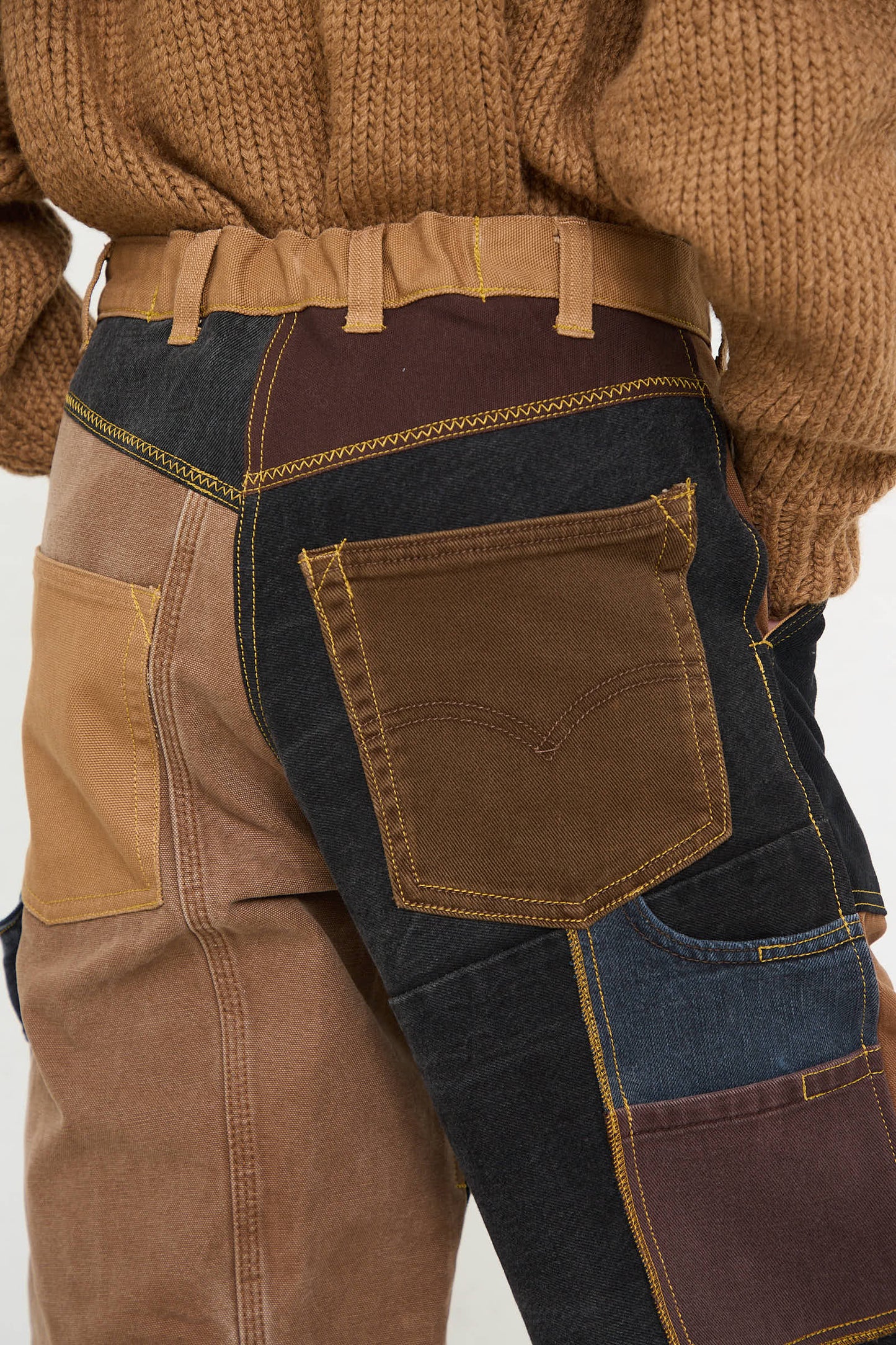 Close-up of a person wearing the Reworked Jeans in Black and Brown I by WildRootz, handmade in New York. These jeans feature recycled denim with various colors and textures. Paired with a brown knit sweater, these pants showcase prominent stitching and a large pocket on the side.