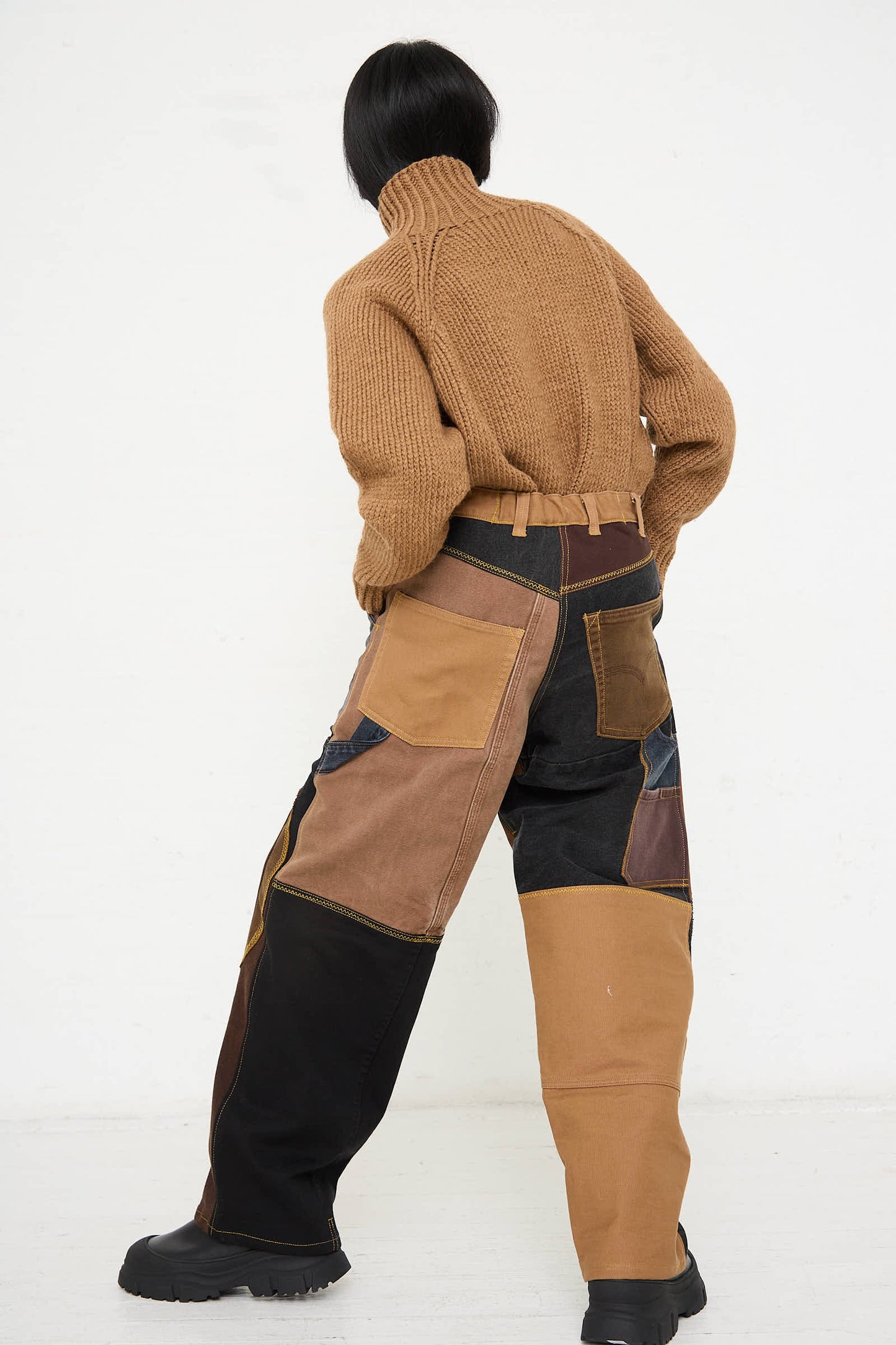 A person with short black hair wears a brown turtleneck sweater and WildRootz's Reworked Jeans in Black and Brown I, featuring various shades of brown and black, standing against a plain white background.