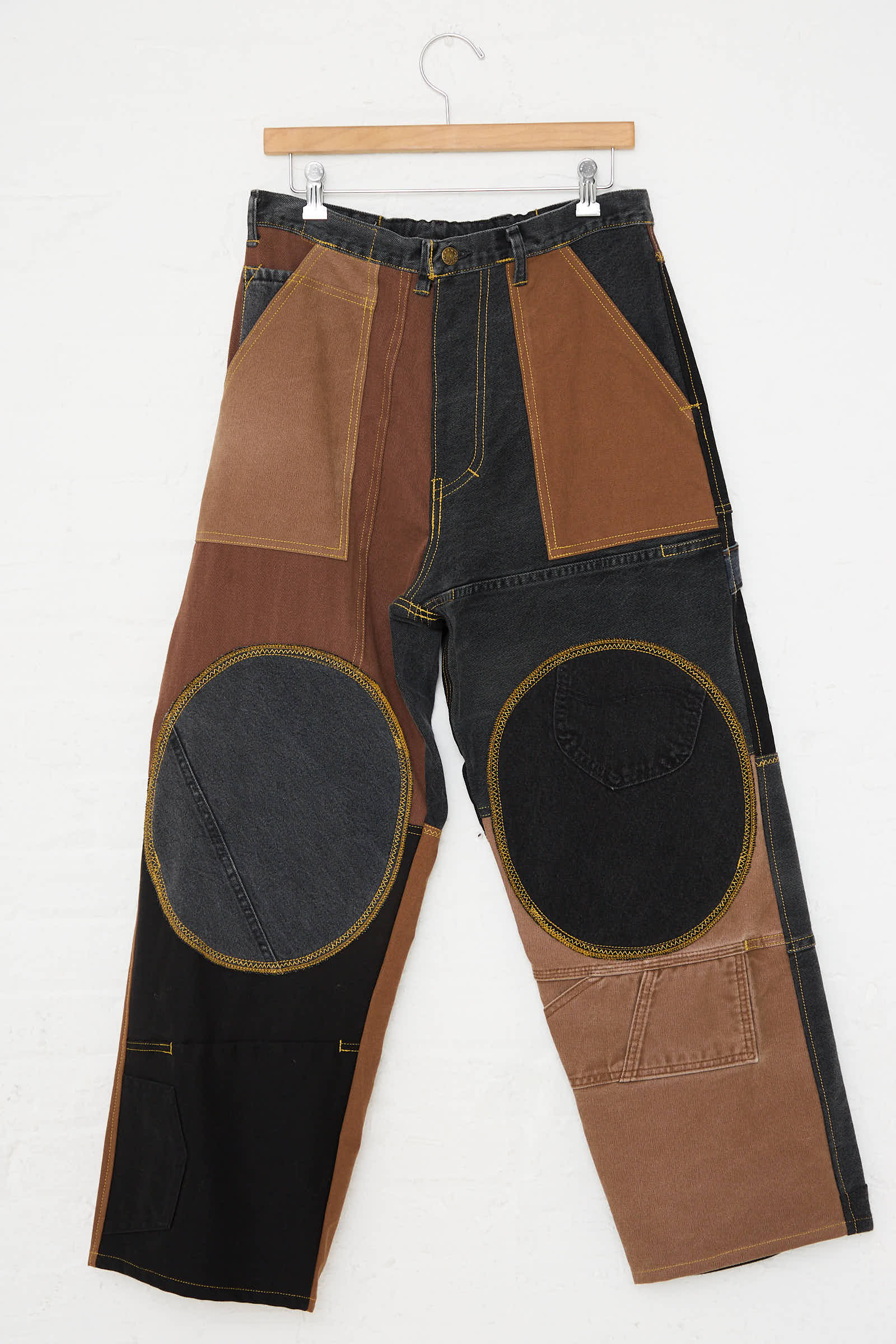 The Reworked Jeans in Black and Brown II by WildRootz, a pair of handmade patchwork jeans crafted from recycled denim, featuring an array of brown, black, and gray patches along with large circular knee patches, elegantly hang on a wooden hanger against a white wall.