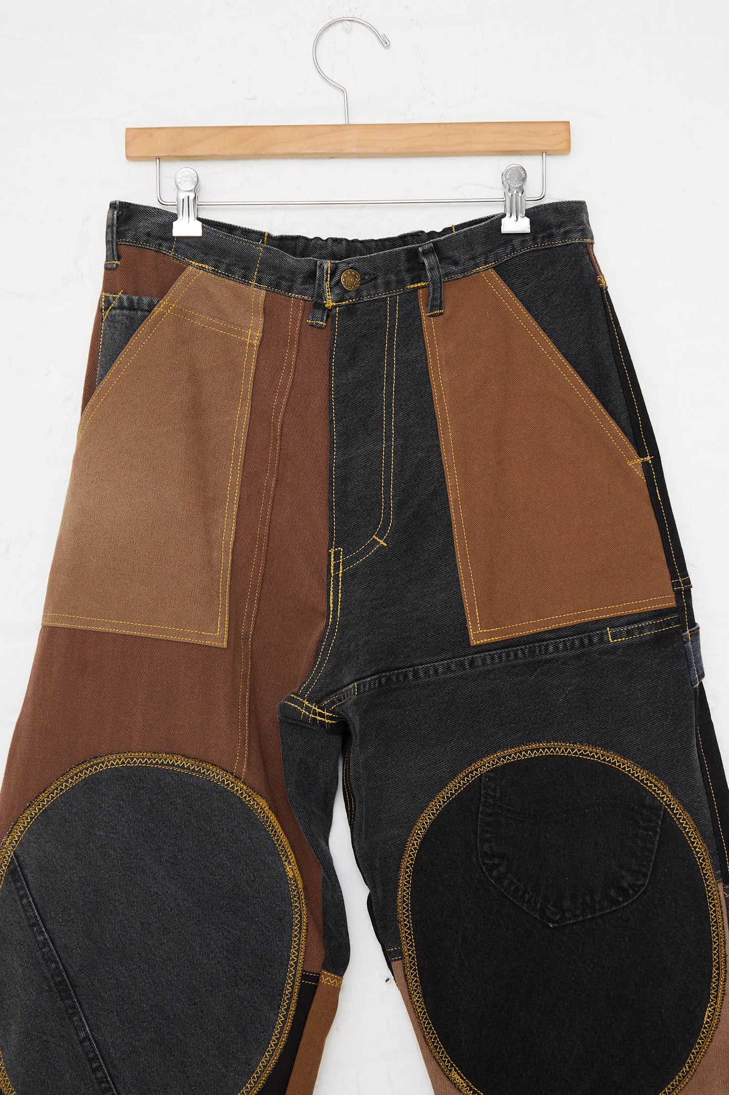 A pair of Reworked Jeans in Black and Brown II by WildRootz, featuring a patchwork design with brown and dark grey fabrics, visible yellow stitching, and large circular patches on the knees, handmade in New York from recycled denim, hanging on a wooden hanger against a white background.