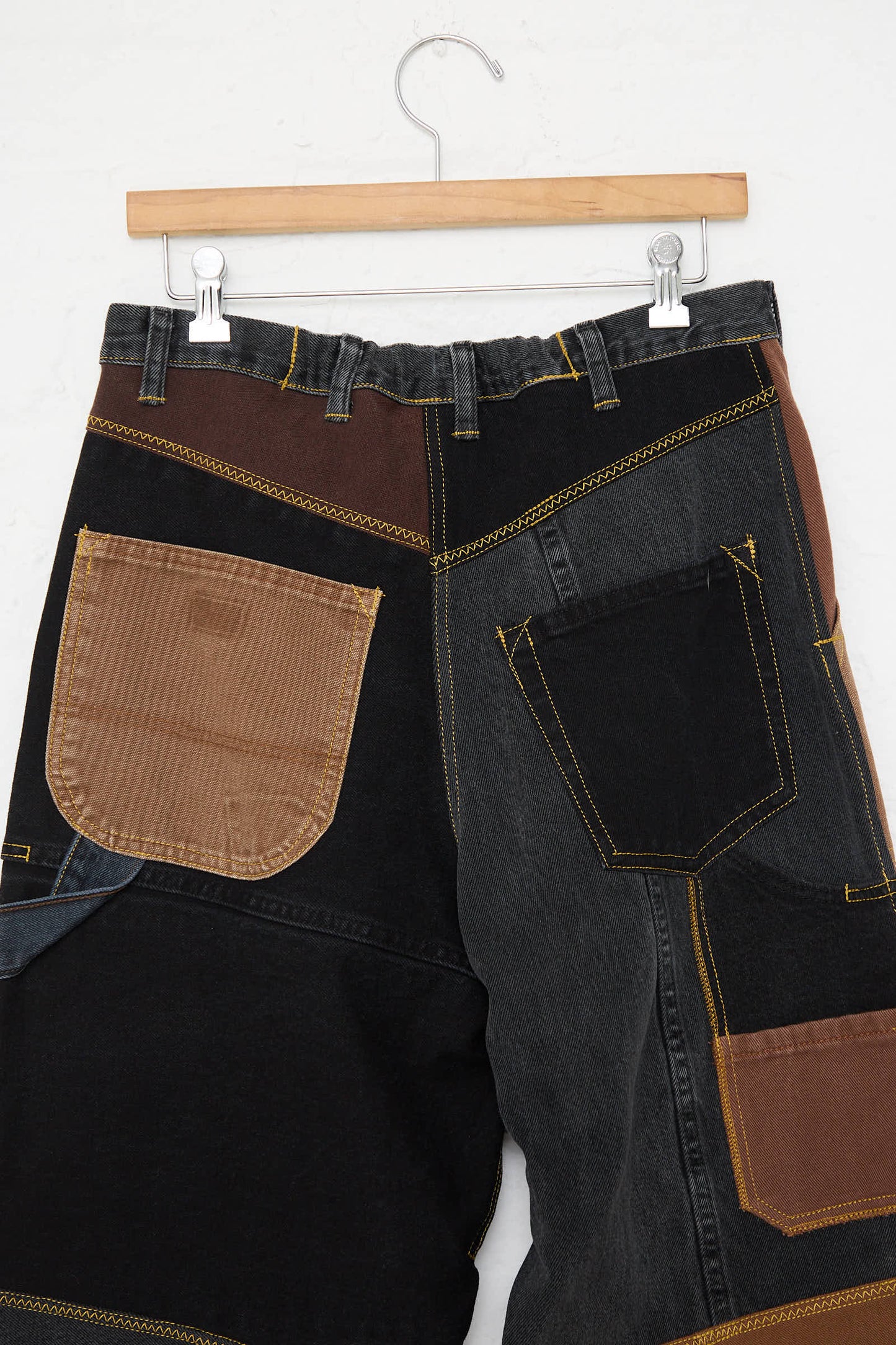 The WildRootz Reworked Jeans in Black and Brown II, handmade in New York, feature a mix of black, brown, and blue recycled fabric patches. Displayed on a wooden hanger against a plain white background.