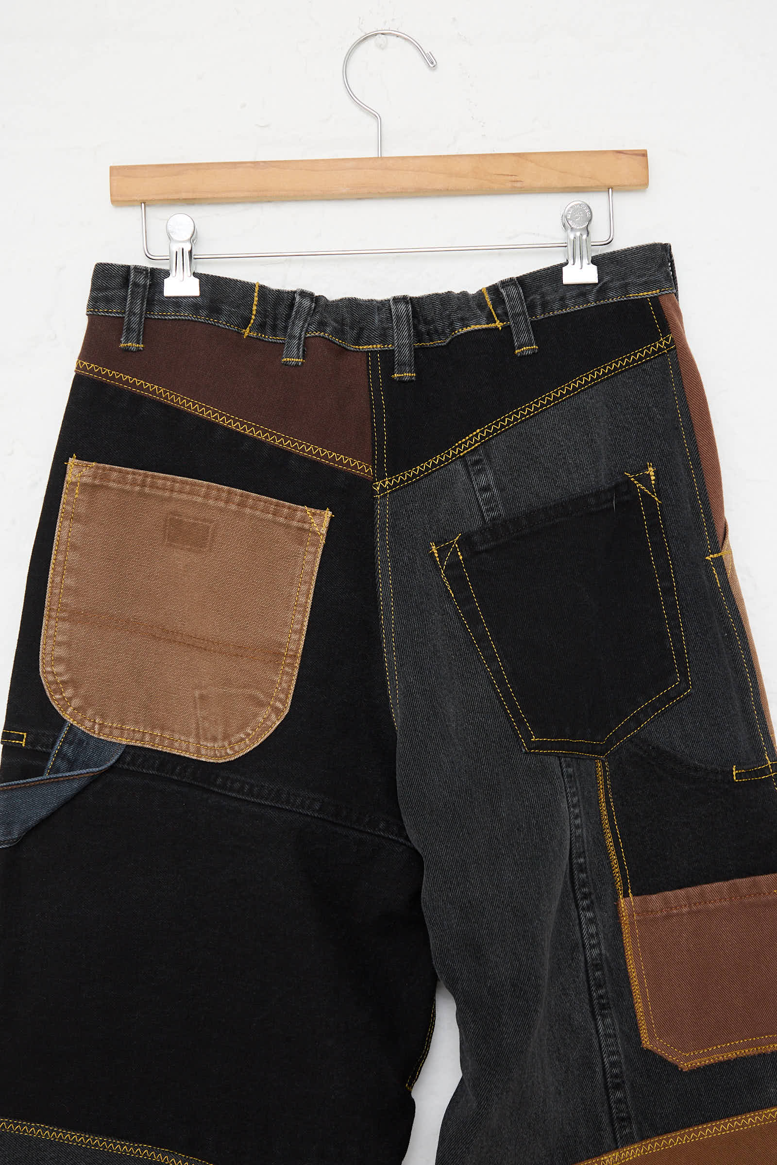 The WildRootz Reworked Jeans in Black and Brown II, handmade in New York, feature a mix of black, brown, and blue recycled fabric patches. Displayed on a wooden hanger against a plain white background.