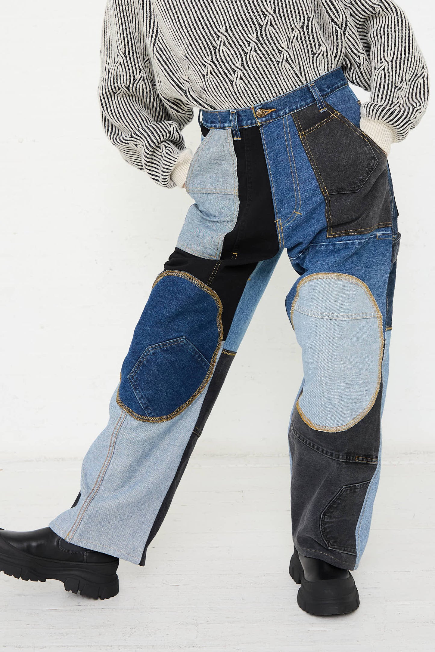 The individual is sporting WildRootz's Reworked Jeans in Blue, Black and Grey I, crafted from recycled denim in an array of shades. They are also wearing a black and white striped sweater while standing in a relaxed pose against a plain, white background.