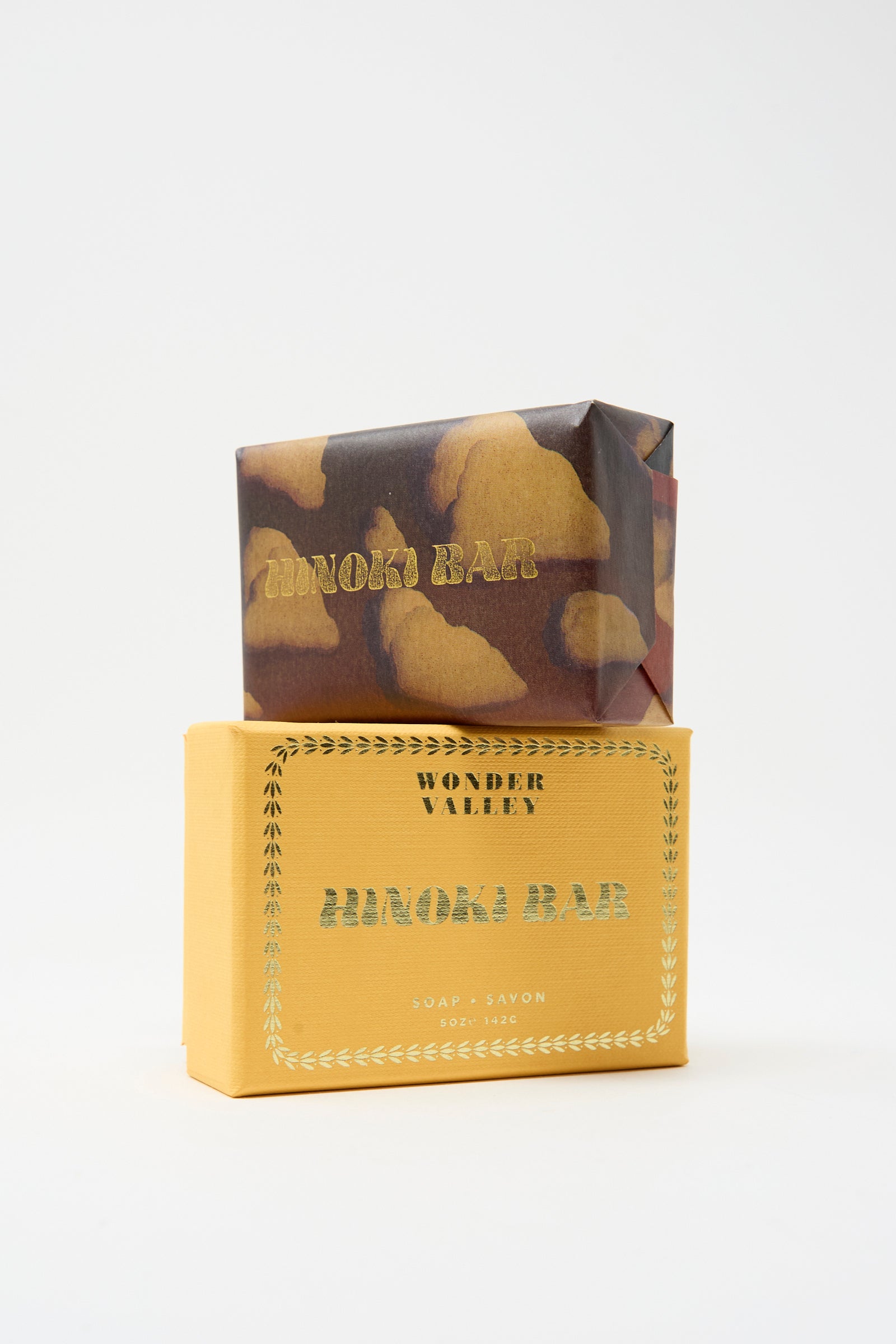 Two rectangular soap bars are stacked; the top is wrapped in brown and yellow paper, while the bottom sits in a yellow box labeled "Wonder Valley Hinoki Bar Soap," a vegan, cruelty-free soap by Wonder Valley.
