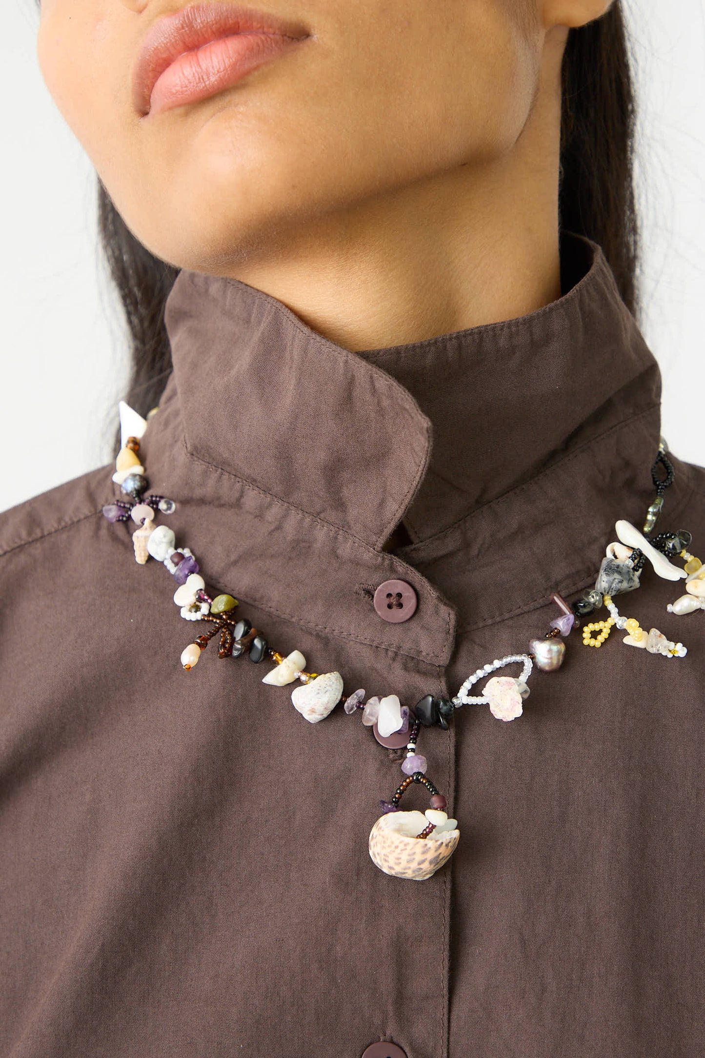 Person wearing a brown button-up shirt, adorned with Yona Kohen's "August is an Angel Necklace," featuring multicolored, irregularly shaped natural stones and a central shell pendant.