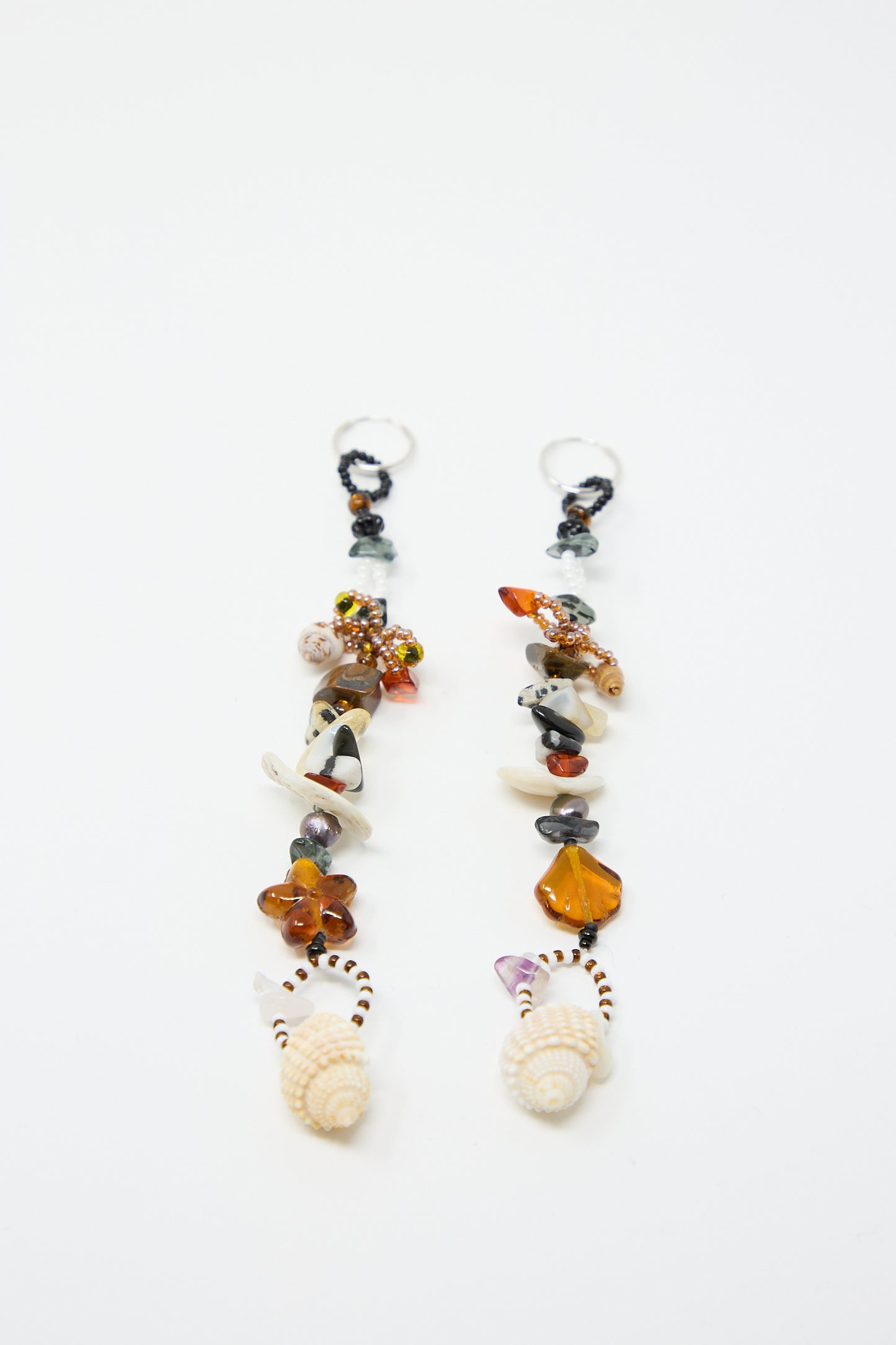 The Coquina Earrings by Yona Kohen are a pair of long, dangling earrings adorned with an eclectic mix of shells, natural stones, and glass beads in various shapes and colors, each ending with a seashell at the bottom, beautifully arranged on a white background.