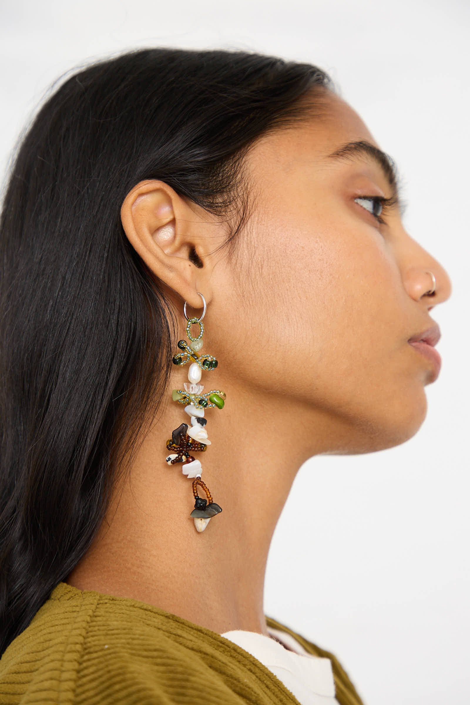 A person with long dark hair is shown in profile, wearing the Yona Kohen Harvest Earrings 2.0, which are large and hand-beaded with various beads and stones. They have a nose ring and are dressed in a green and white outfit.