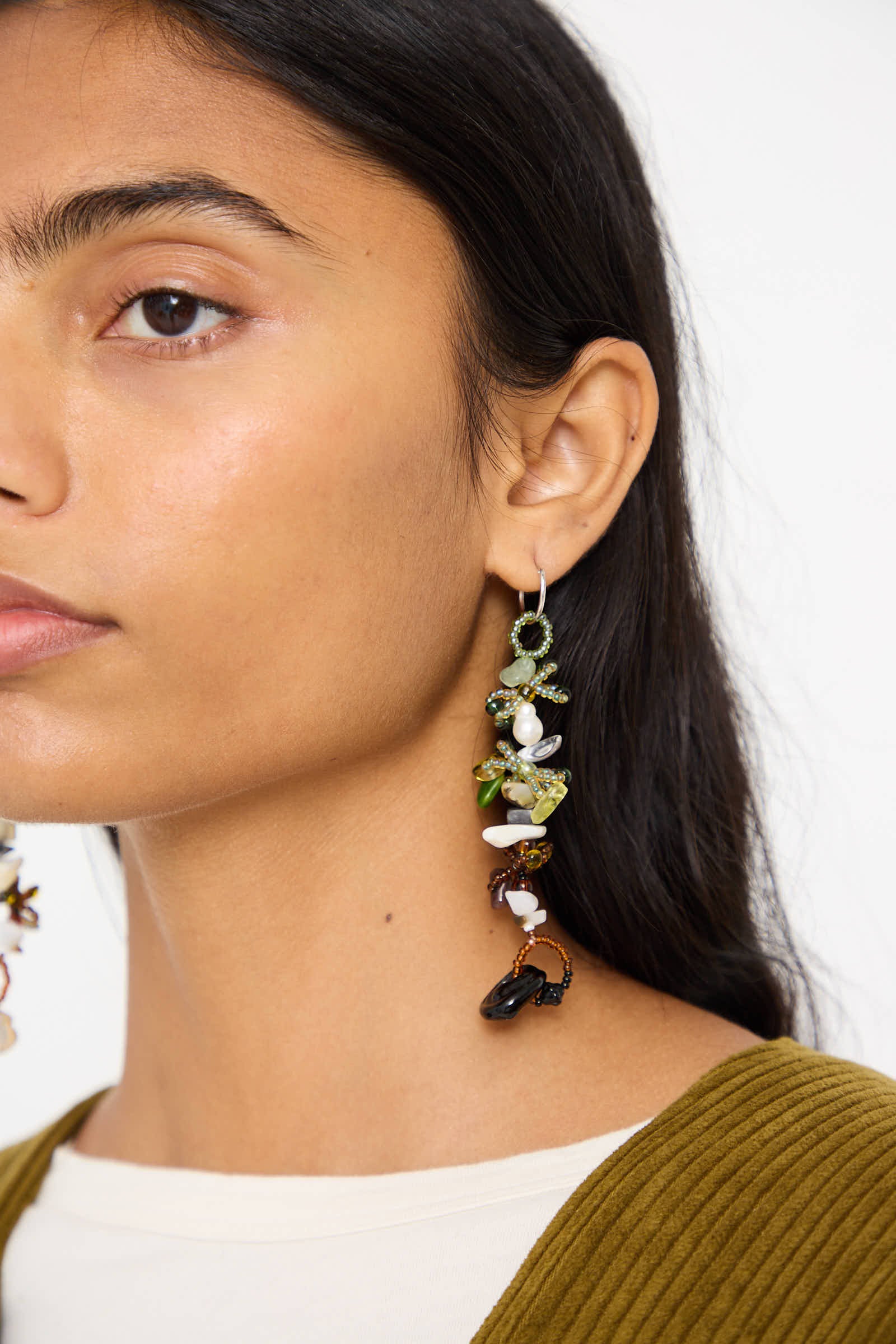 Close-up of a person with long dark hair wearing a green top and detailed, multi-colored hand-beaded Harvest Earrings 2.0 by Yona Kohen, featuring various glass beads and shapes.