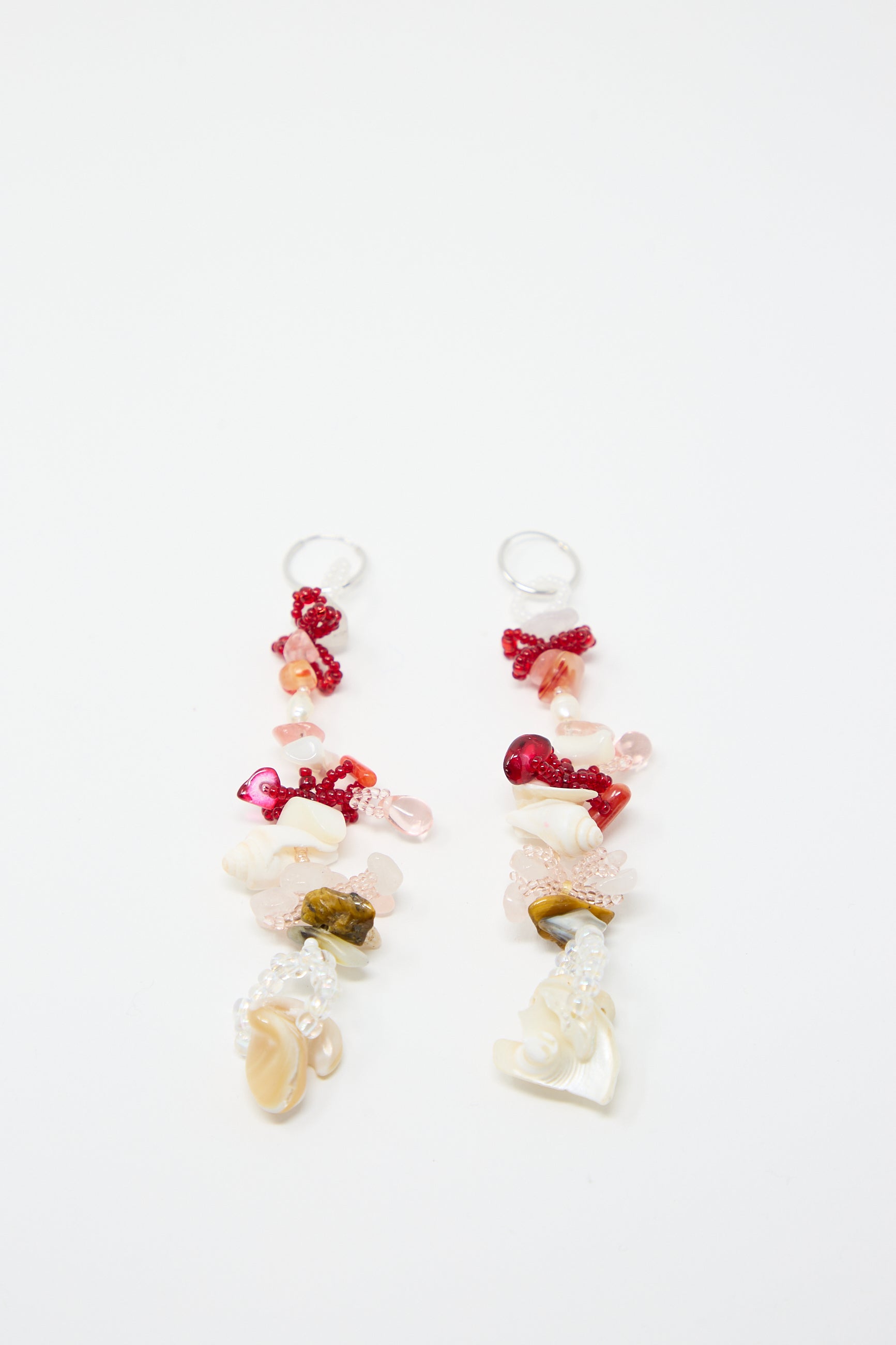 Introducing the Kiraz Earrings by Yona Kohen: a stunning pair of long, dangling hand-beaded earrings featuring an assortment of beads and stones in shades of pink, red, and cream, arranged in a whimsical and eclectic design on sterling silver hoops.