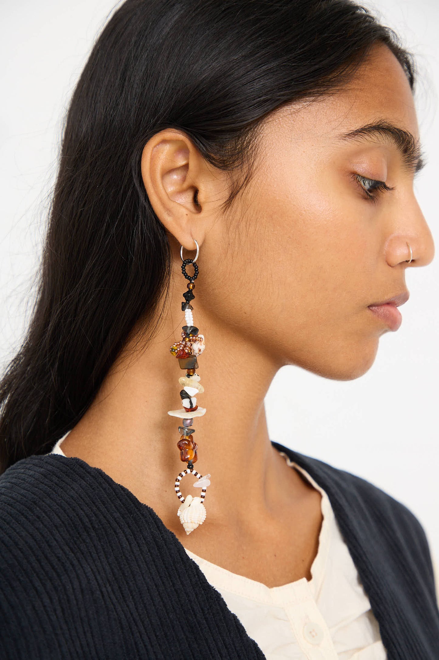 A person with long dark hair wearing a black cardigan and a white shirt showcases the Yona Kohen Coquina Earrings, which feature various beads and ornaments crafted from sterling silver. The background is plain white.