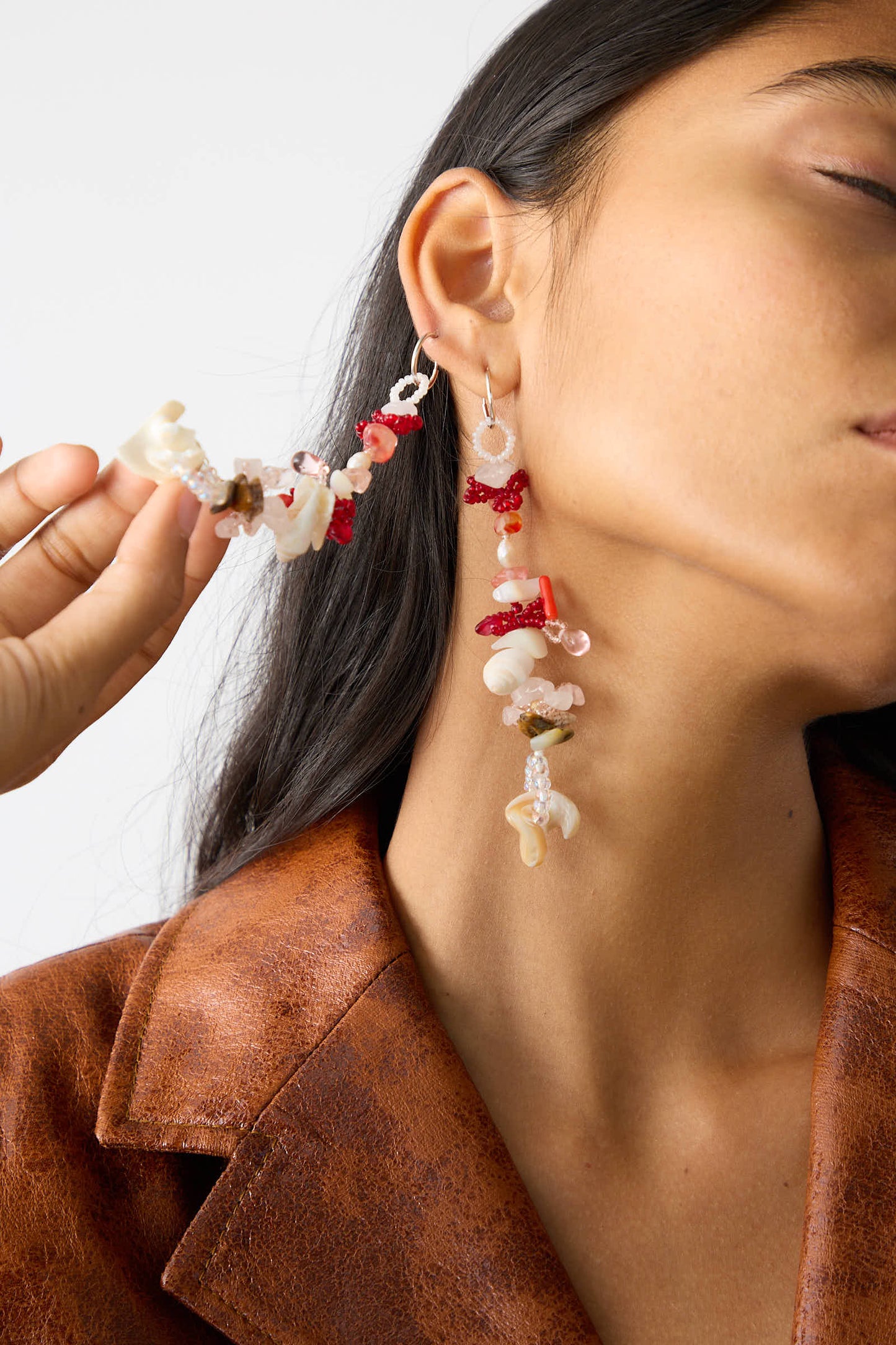 A person with long hair wearing a brown jacket showcases the intricate Kiraz Earrings by Yona Kohen, featuring various colored beads and stones. They hold one earring near their ear, highlighting the delicate design adorned with glass beads.