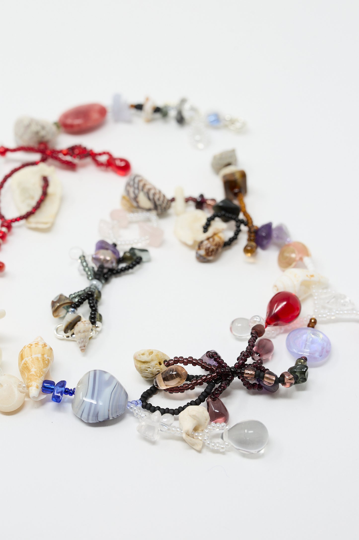 The Love is Stronger Than Pride Necklace by Yona Kohen showcases a range of hand-beaded necklaces adorned with assorted glass beads and semi-precious stones, including vibrant red, purple, and blue beads, all elegantly displayed on a white surface.