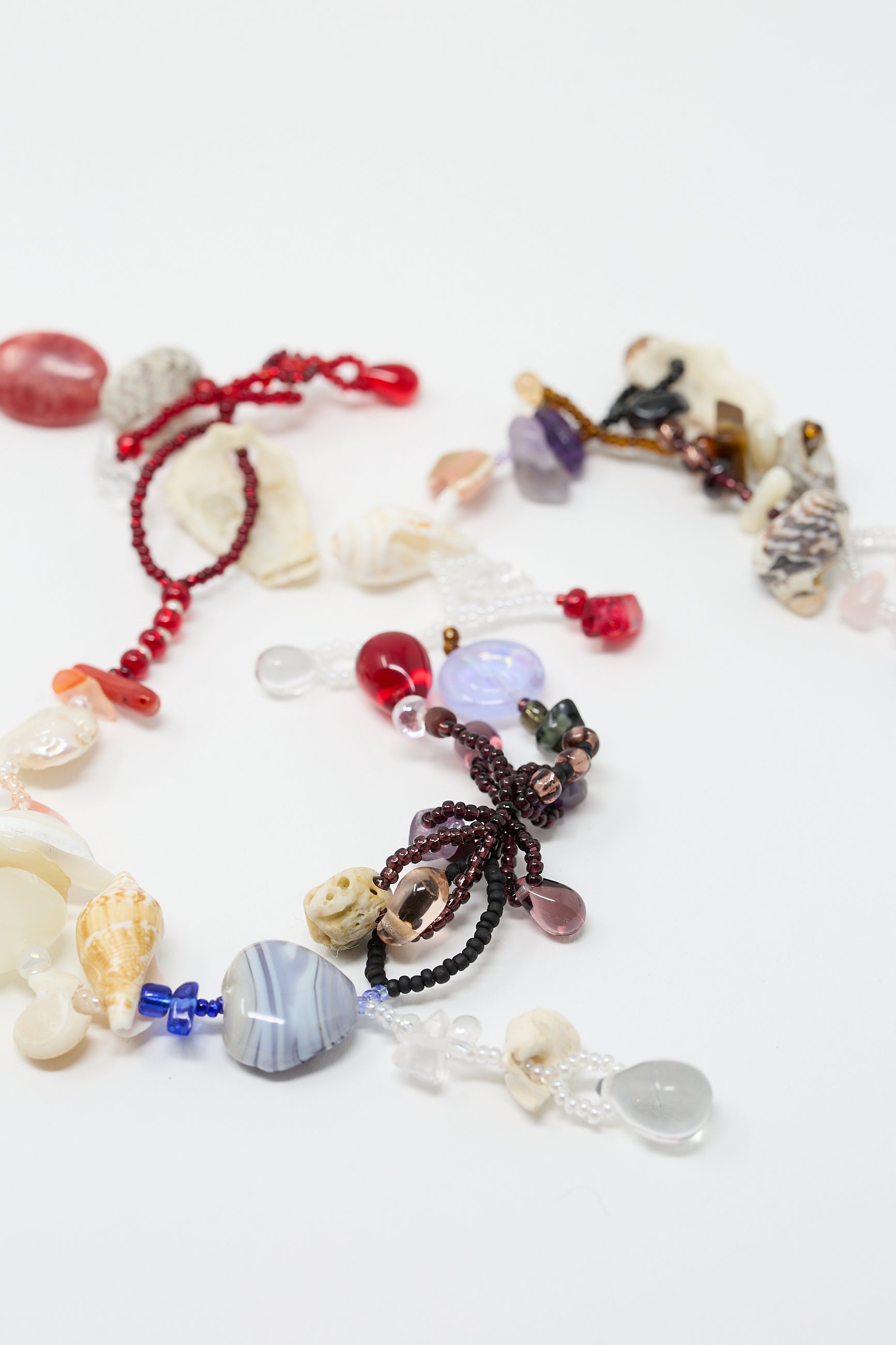 Assorted beads and glass beads in various shapes, sizes, and colors are arranged on a white surface, including heart, shell, and flower-shaped beads in red, blue, purple, and white hues for the "Love is Stronger Than Pride Necklace" by Yona Kohen.