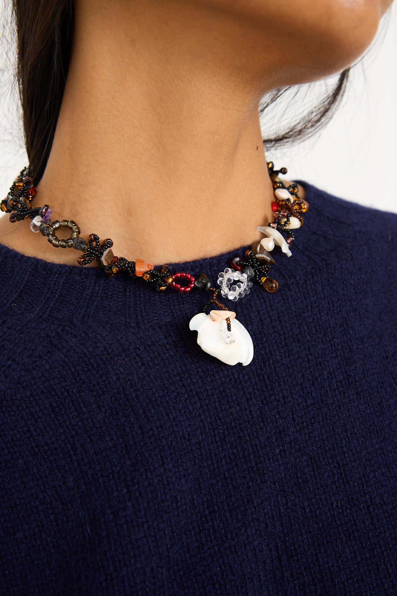 A person wearing a dark blue sweater adorned with the Yona Kohen Silkworm Necklace in Black, featuring colorful hand-beaded details with various shapes, semi-precious stones, and a white decorative piece at the center.