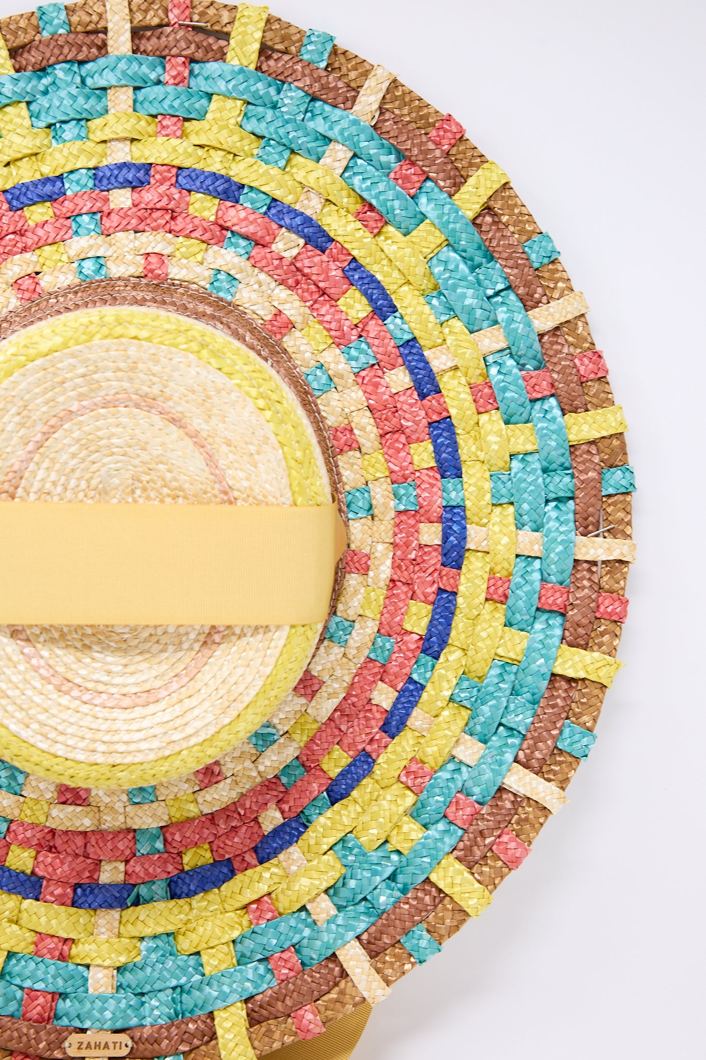 Close-up of a colorful handmade Straw Cuchi Tris-Tras No. 94 Hat with Laces with concentric patterns in yellow, blue, red, and green, featuring a yellow headband. The wide brim is crafted from braided straw. The brand name "Zahati" is visible on the bottom edge of the hat.