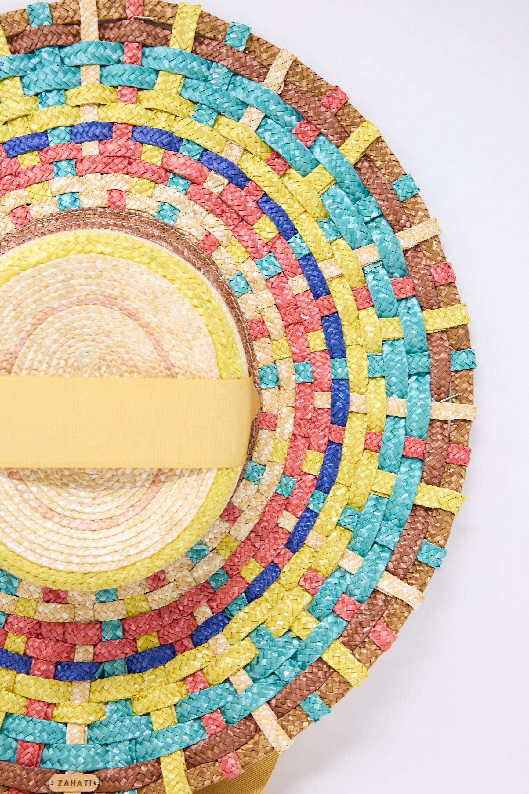 Close-up of a colorful handmade Straw Cuchi Tris-Tras No. 94 Hat with Laces with concentric patterns in yellow, blue, red, and green, featuring a yellow headband. The wide brim is crafted from braided straw. The brand name "Zahati" is visible on the bottom edge of the hat.