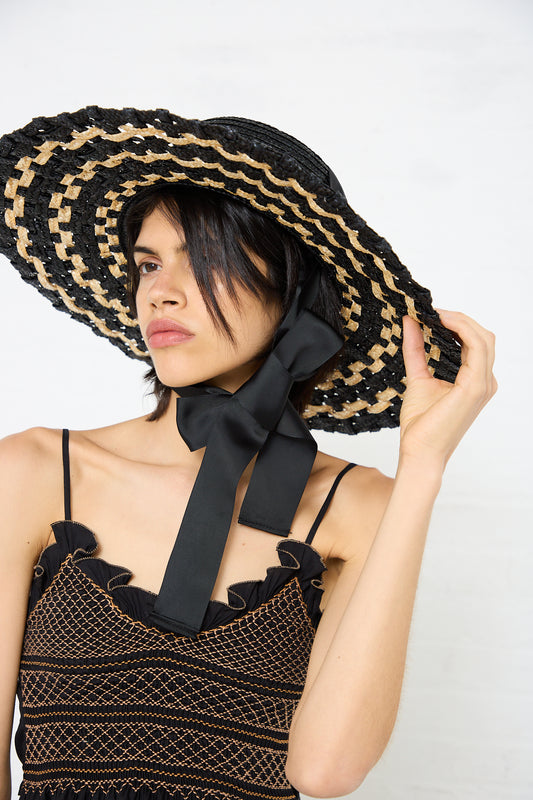 A woman wearing a Zahati Straw Cuchi Tris-Tras No. 104 Hat with Laces, holding the wide brim with one hand, and dressed in a black and brown patterned spaghetti strap top.
