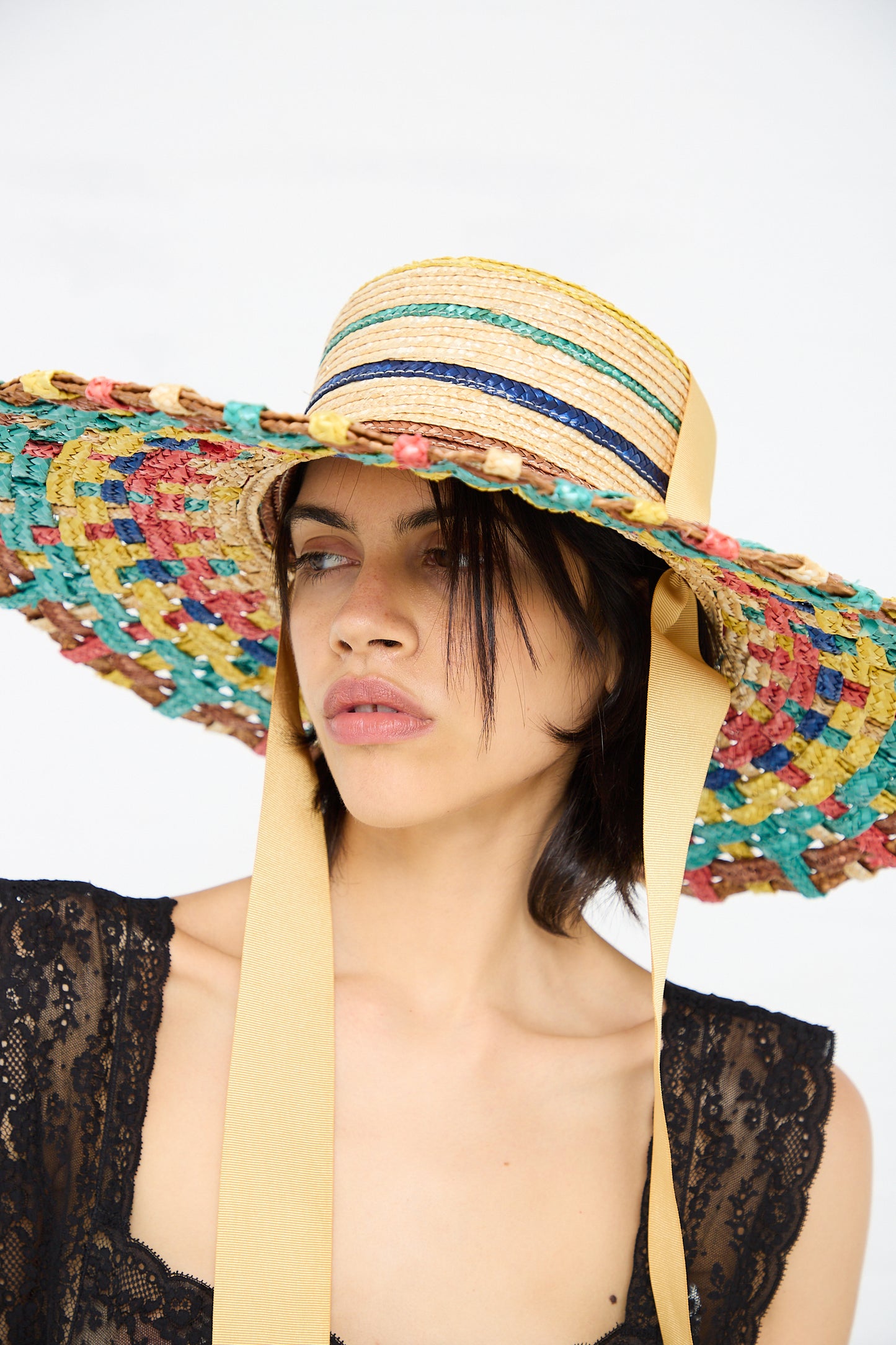 A woman wearing a colorful, wide-brimmed Zahati Straw Cuchi Tris-Tras No. 94 Hat with Laces with a braided straw design and beige ribbon, dressed in a sheer black lace top, looks to the side.
