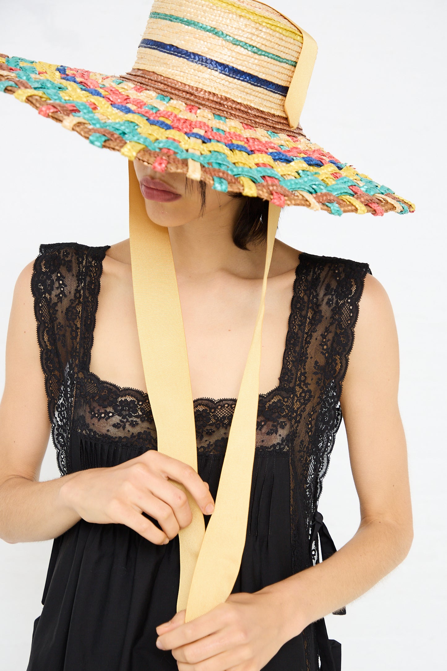 A person wearing a wide-brimmed, multicolored Straw Cuchi Tris-Tras No. 94 Hat with Laces by Zahati and a black lace-trimmed dress.