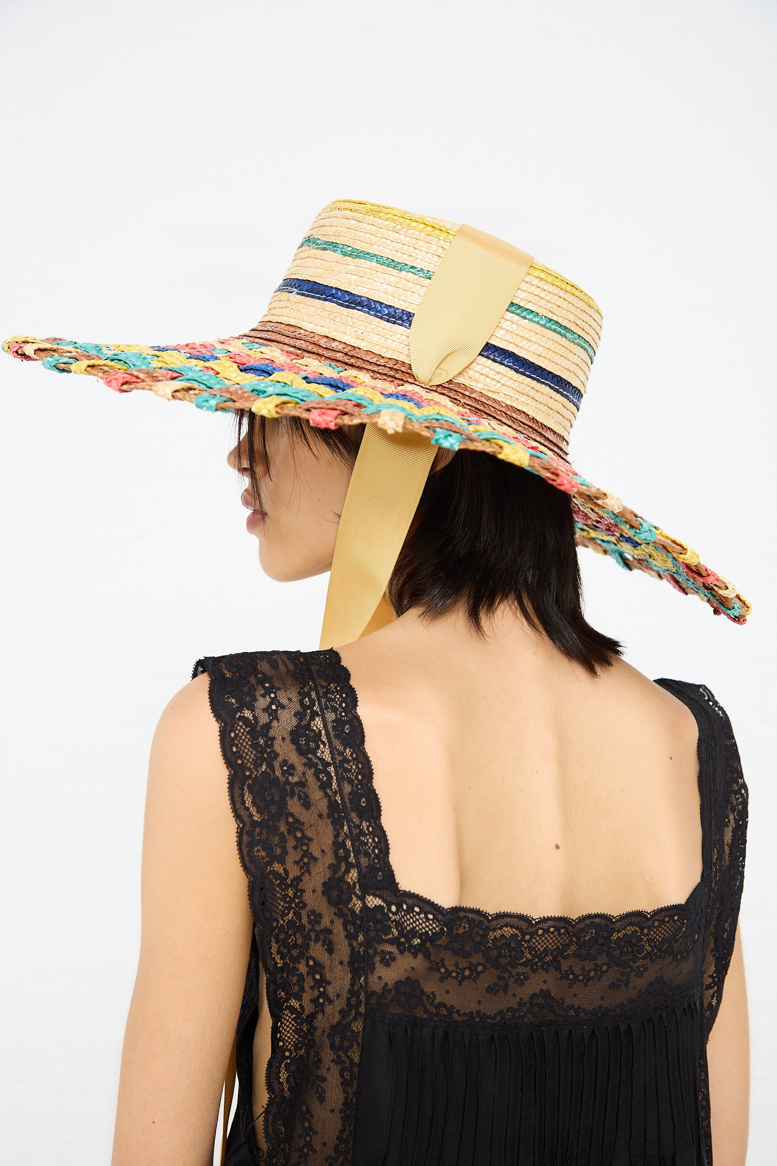 A woman with short dark hair is shown from the side wearing a handmade sun hat with a wide brim and a lace black top. The multicolored Zahati Straw Cuchi Tris-Tras No. 94 Hat with Laces has a yellow ribbon hanging from it.