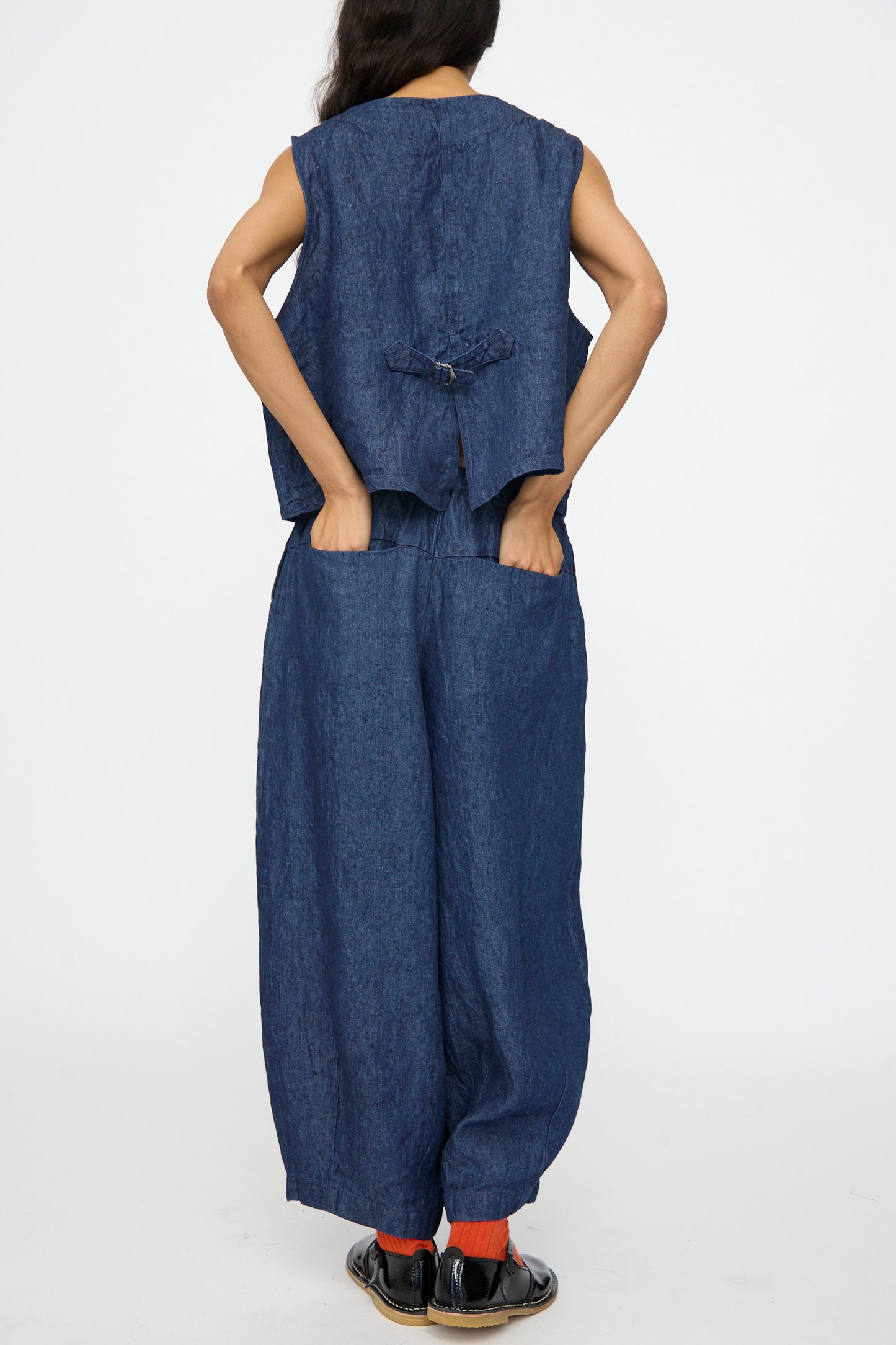 A person faces away, wearing a sleeveless blue denim top, nest Robe 10.oz Linen Denim Wide Tapered Trousers in Indigo Blue, black shoes, and orange socks, all on a plain white background.