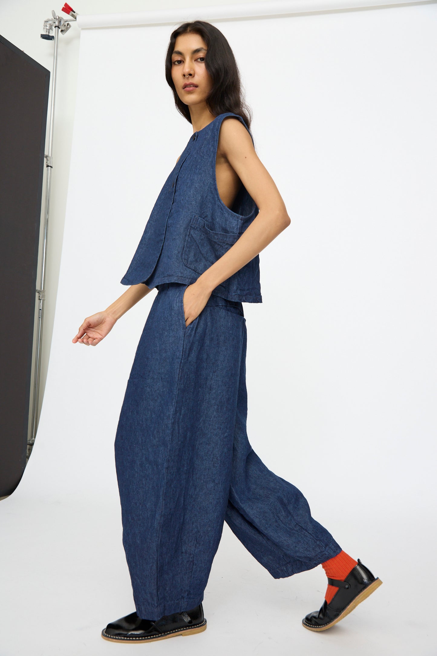 A person poses in a studio wearing the nest Robe 10.oz Linen Denim Wide Tapered Trousers in Indigo Blue with a sleeveless denim top, black shoes, and orange socks against a plain white background for a relaxed yet stylish look.