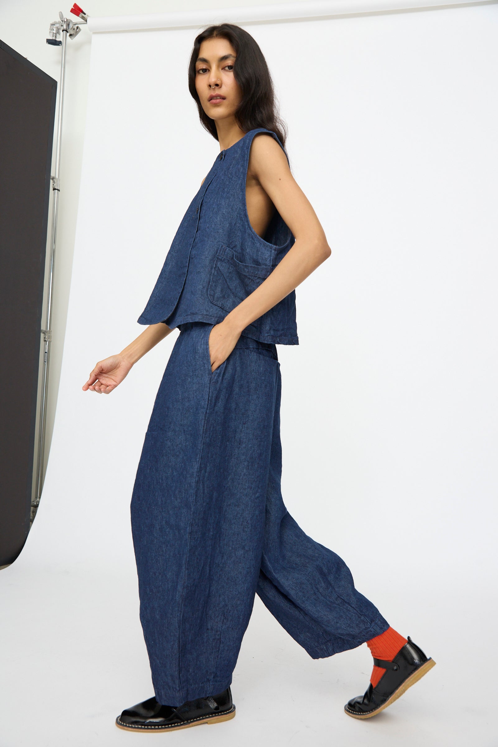 A person poses in a studio wearing the nest Robe 10.oz Linen Denim Wide Tapered Trousers in Indigo Blue with a sleeveless denim top, black shoes, and orange socks against a plain white background for a relaxed yet stylish look.
