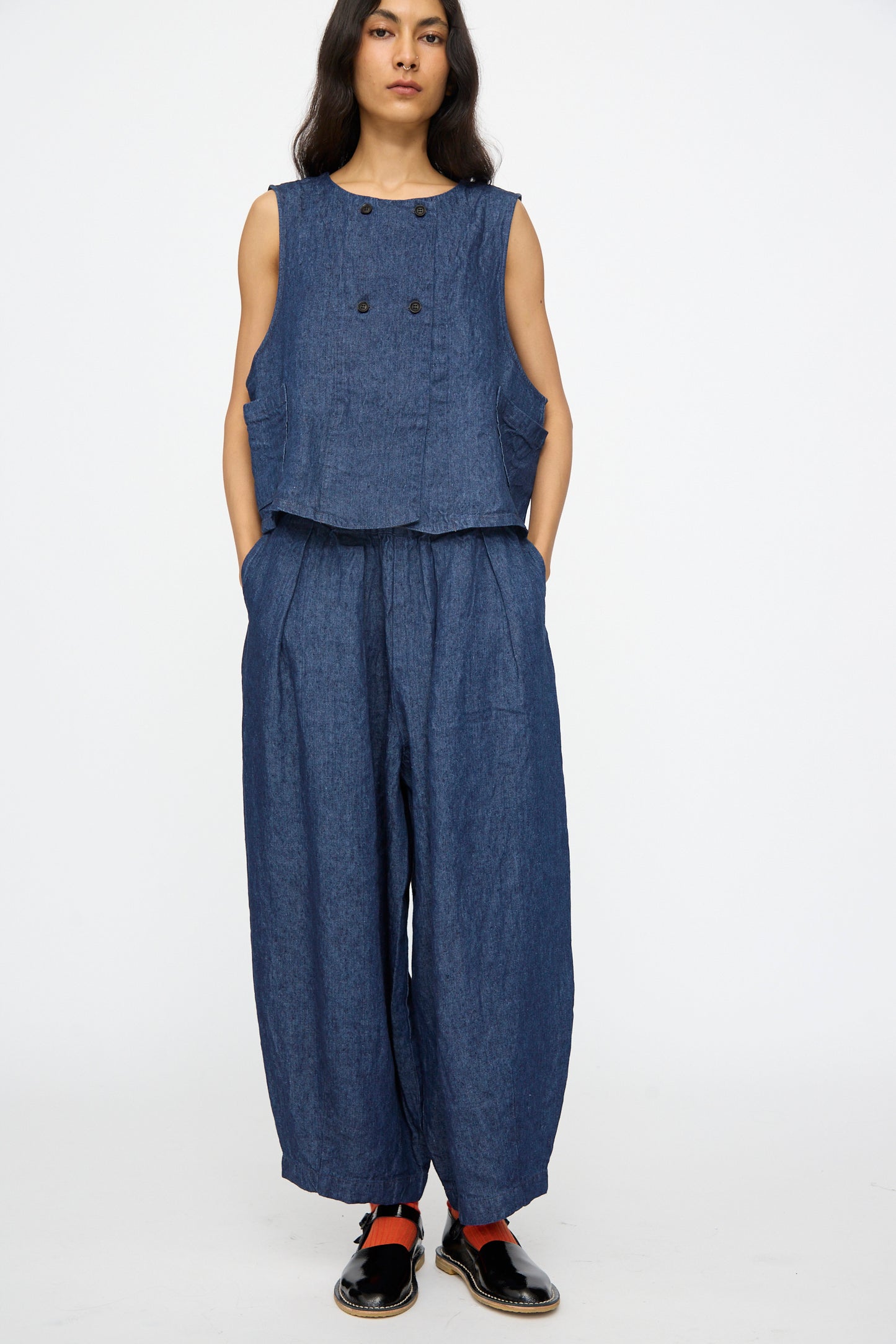 A person stands against a white background wearing nest Robe’s 10.oz Linen Denim Wide Tapered Trousers in Indigo Blue, paired with a sleeveless blue denim top, black shoes, and red socks.