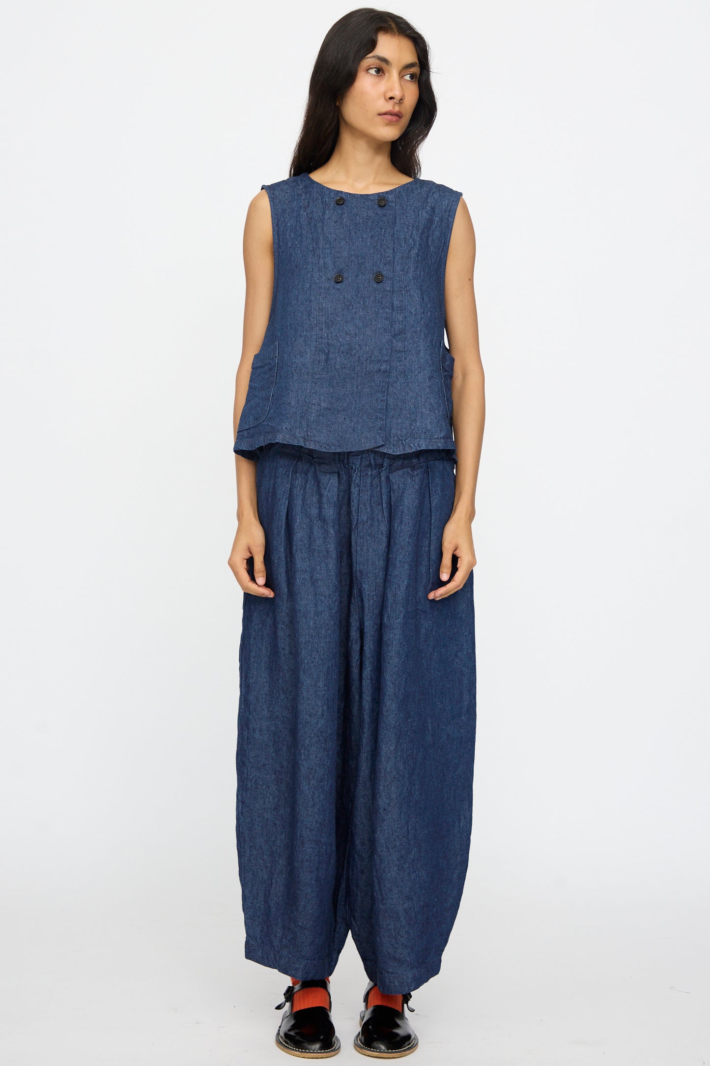 A woman stands facing forward in the nest Robe 10oz. Linen Denim Vest in Indigo Blue with matching loose blue pants and black shoes, set against a plain white background. Authentic craftsmanship, made in Japan.