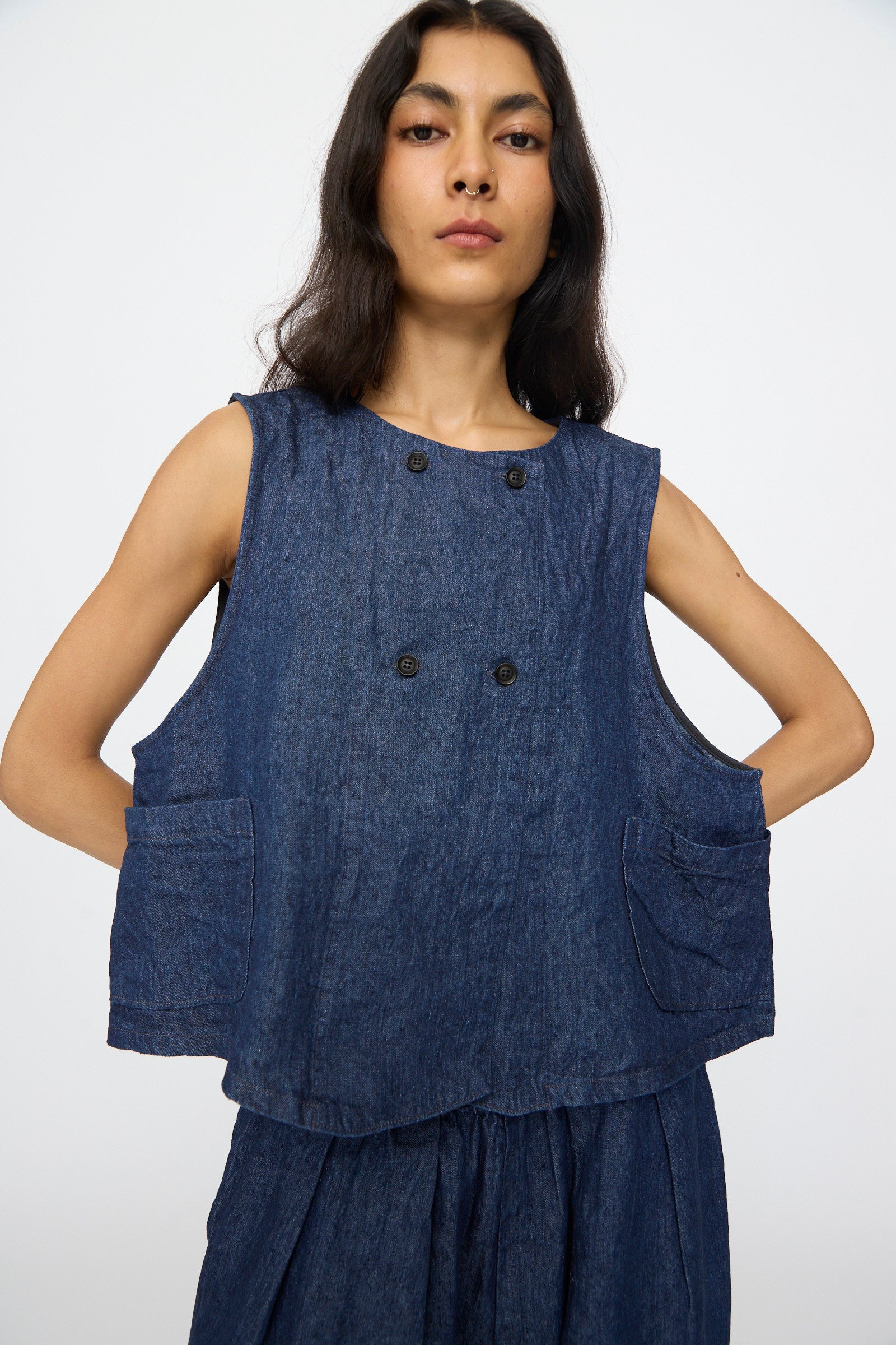 A person wears the nest Robe 10oz. Linen Denim Vest in Indigo Blue, featuring black buttons and two front pockets, paired with matching blue bottoms against a plain light background. The boxy vest lends a modern edge to the outfit.
