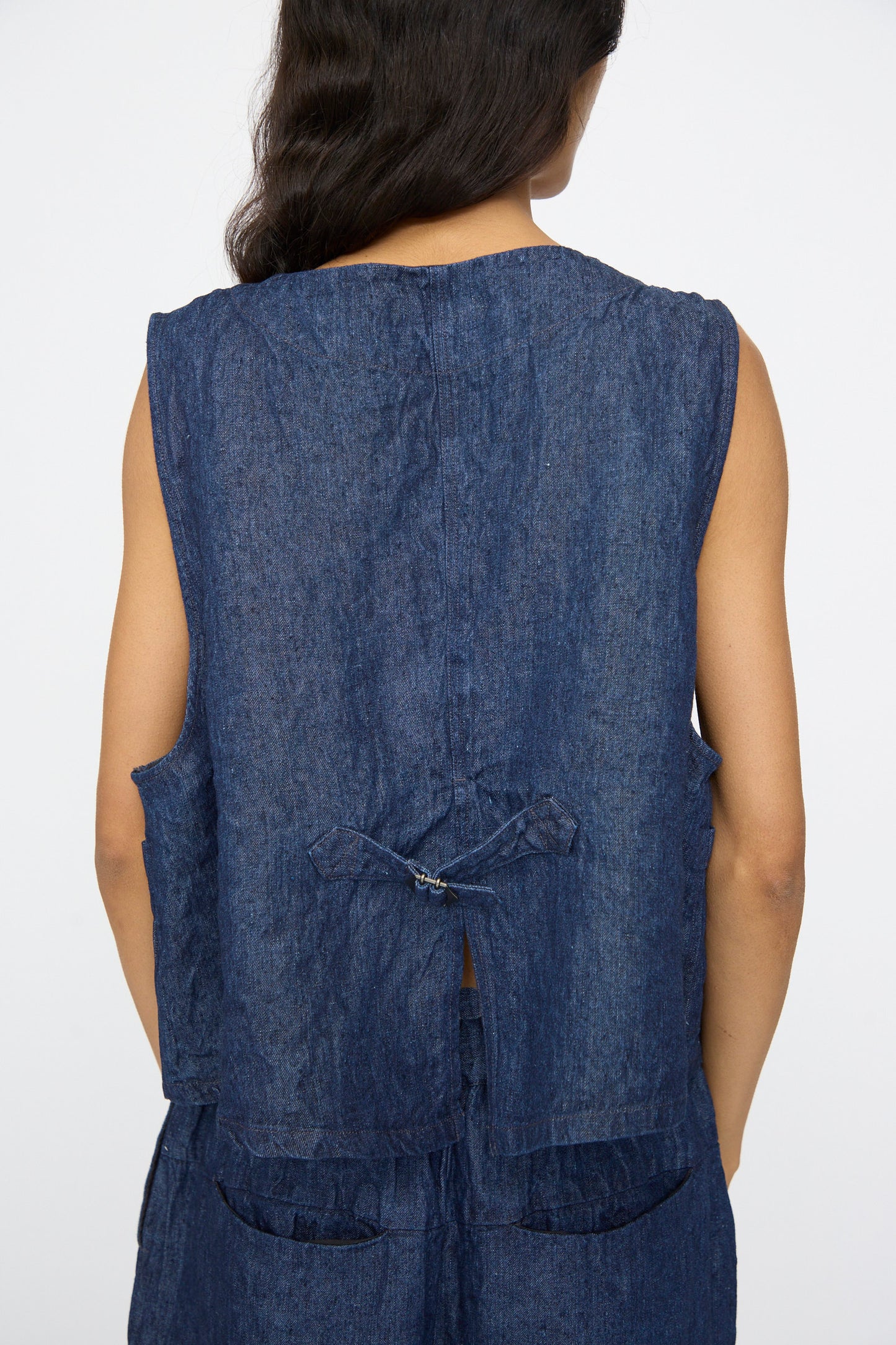 A person with long dark hair is shown from behind wearing the nest Robe 10oz. Linen Denim Vest in Indigo Blue, made in Japan, featuring a boxy fit, back tie detail, and matching bottoms.